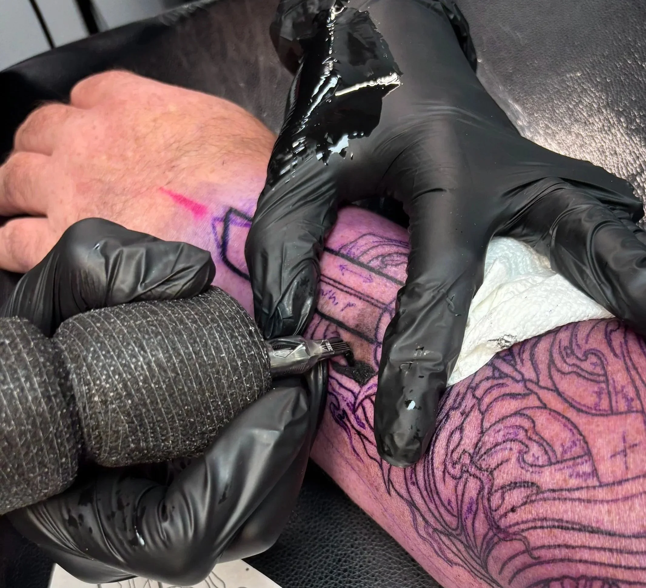 A tattoo artist is tattooing a design onto a person's skin, with the person's hand visible and black gloves on the artist's hands. The tattoo design appears to include some lines and shapes.