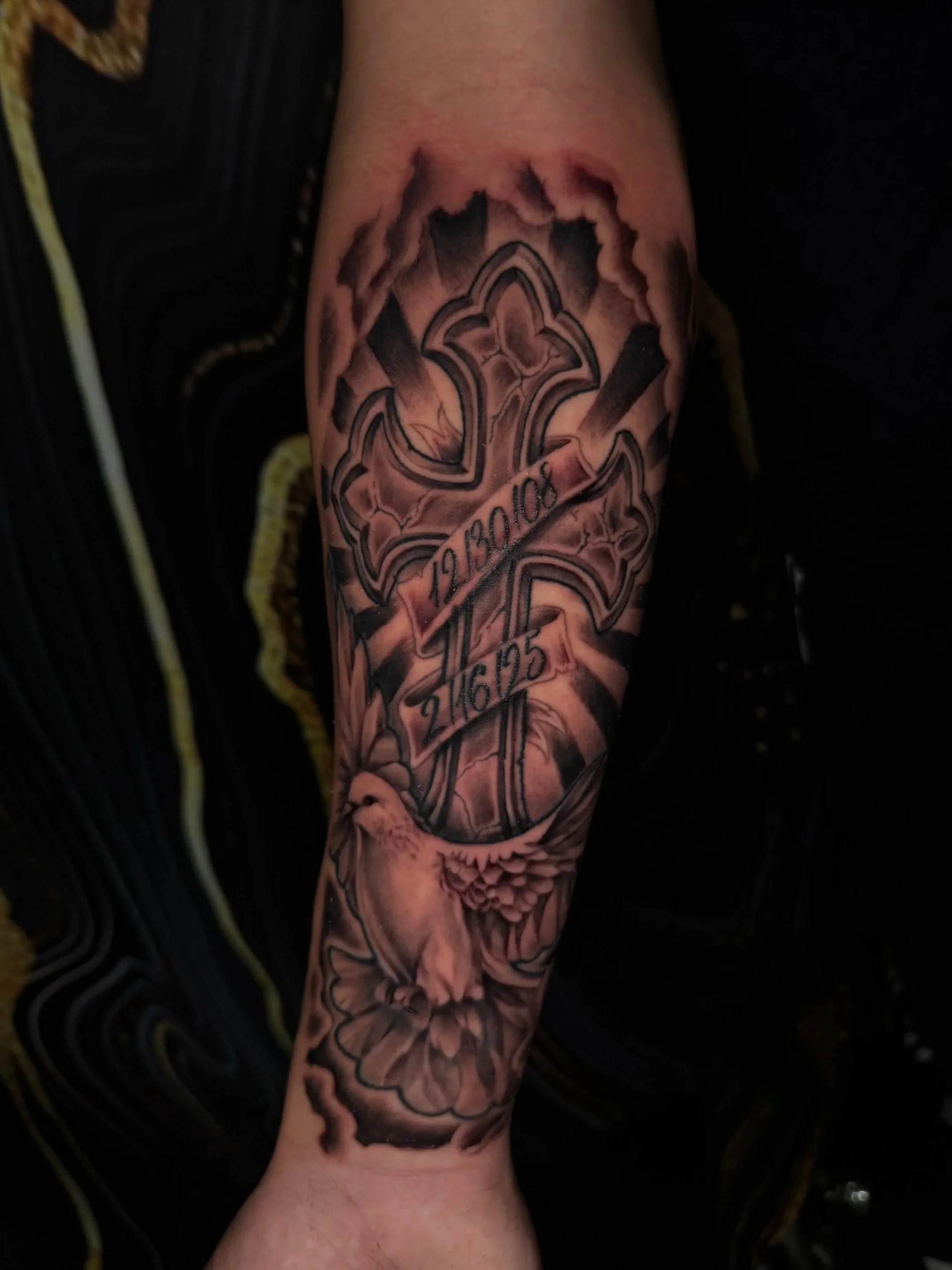 Tattoo of a cross with black shading and a ribbon with dates on a person's arm.