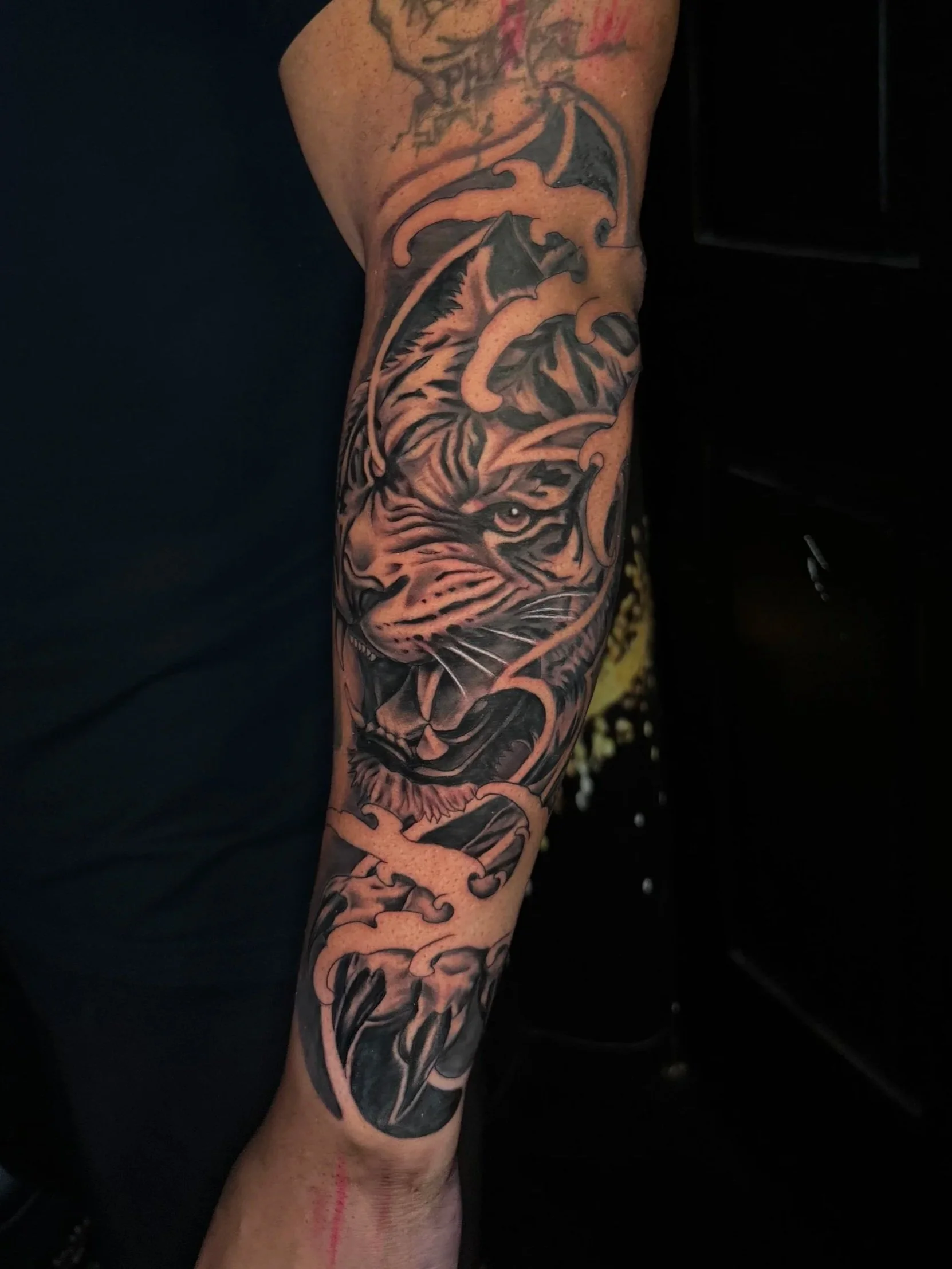 A detailed black and grey tattoo on a person's arm, featuring a fierce tiger's face with sharp eyes and fur markings, intertwined with decorative swirling patterns and abstract elements.