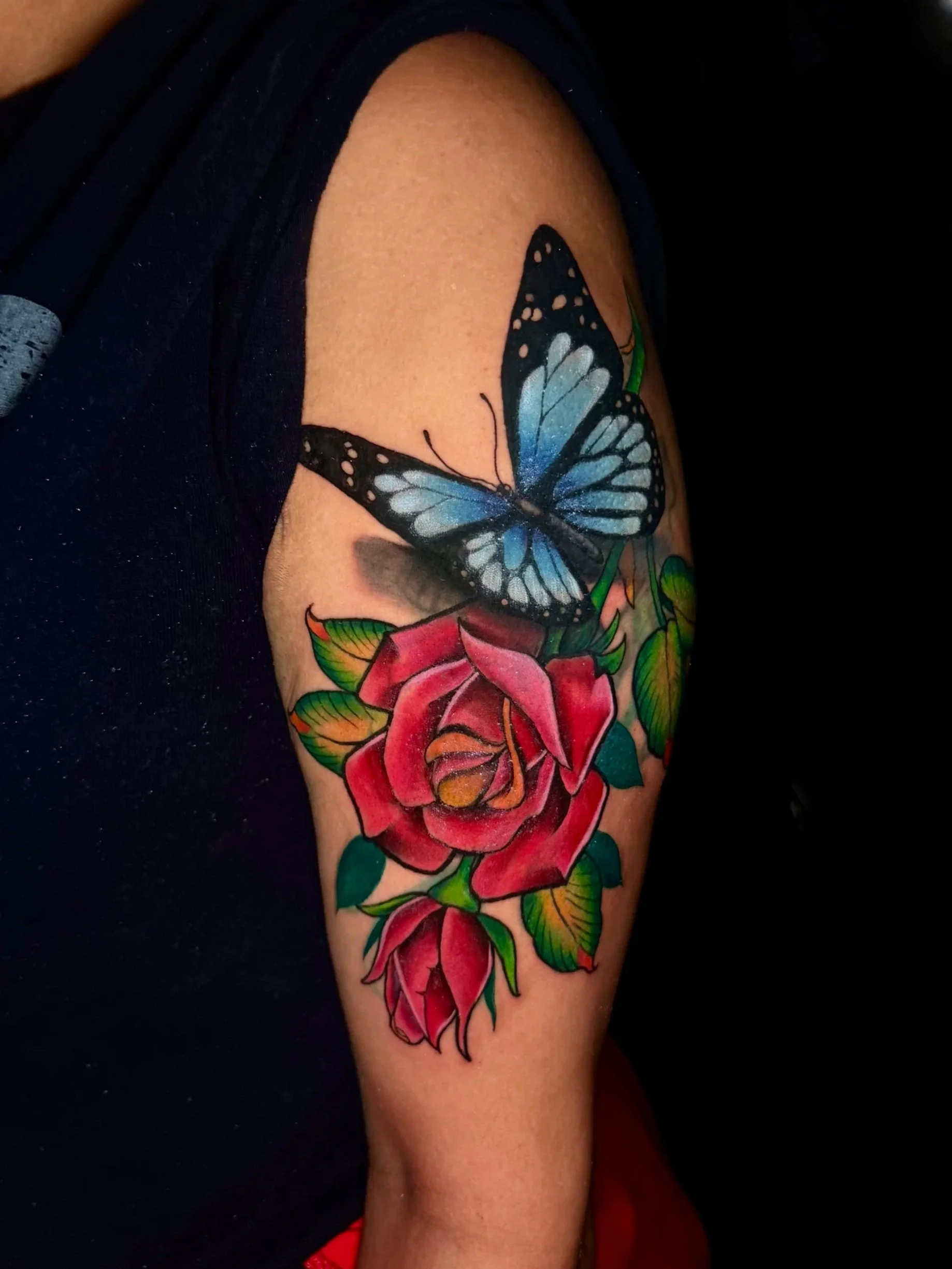 A colorful tattoo of a blue butterfly with black and white spots on the wings, perched on a vibrant red rose with green leaves, on a person's upper arm.