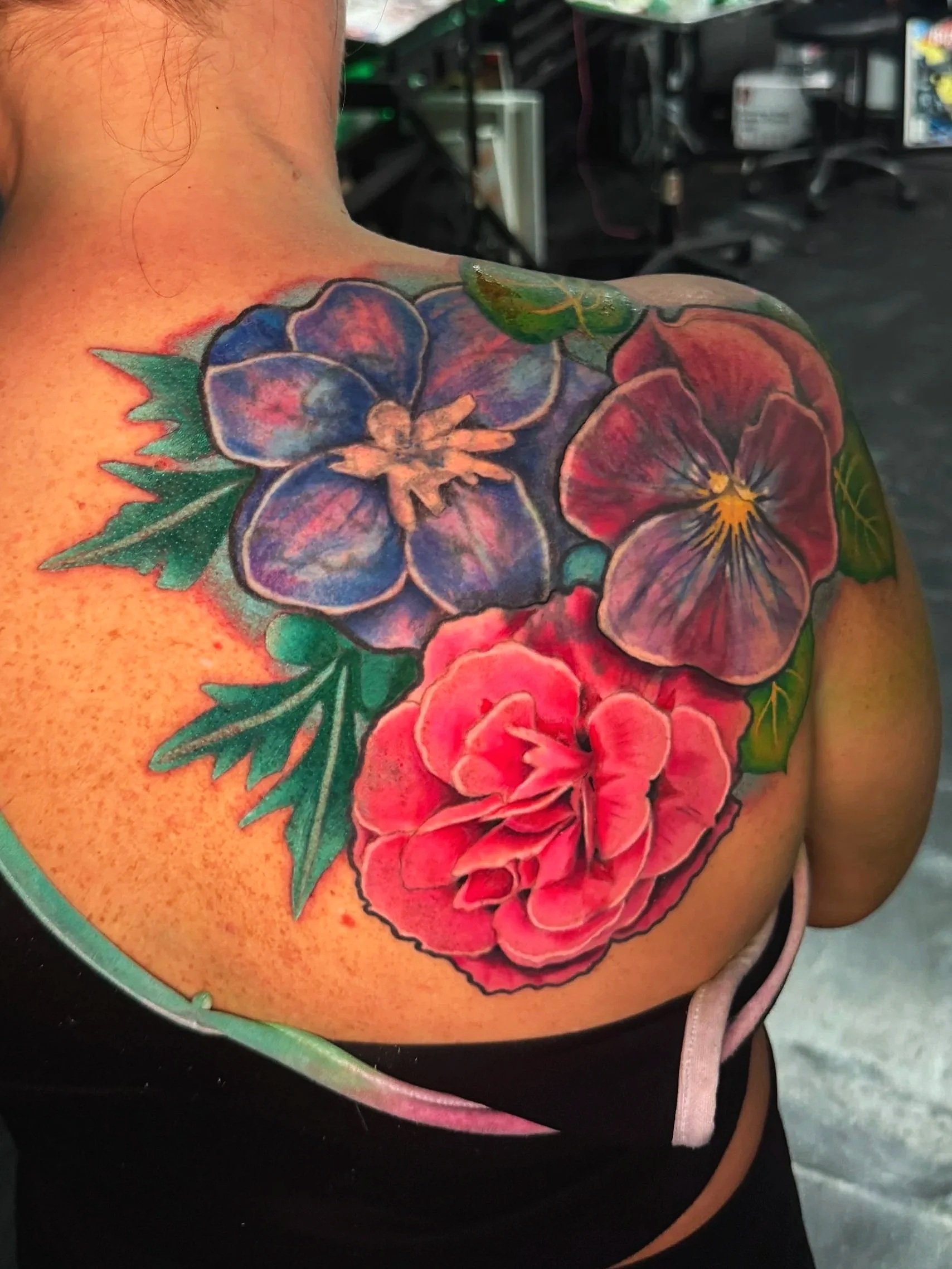 Colorful tattoo of blue and red flowers with green leaves on a person's shoulder.