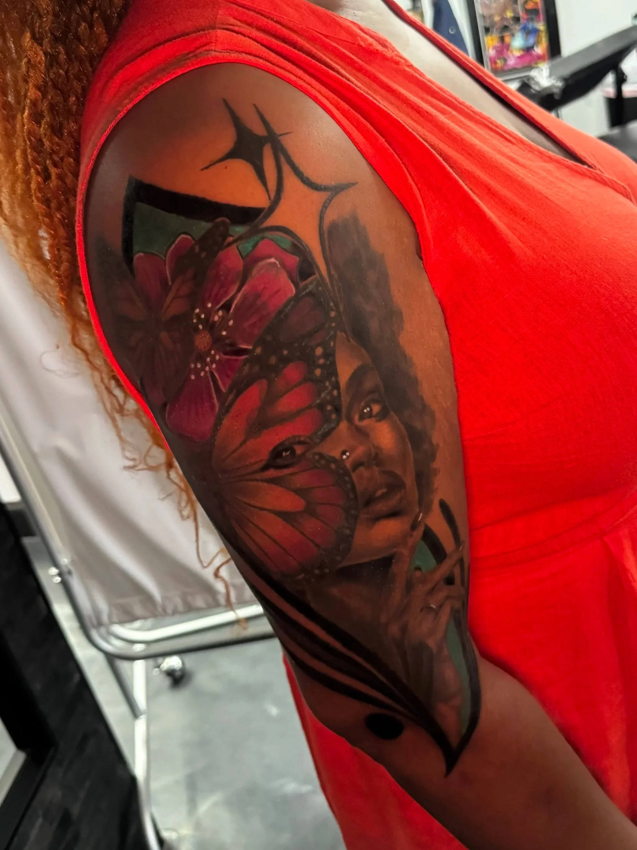 A tattoo of a butterfly and a flower on a person's upper arm, with part of the shoulder and arm visible, wearing a red shirt.