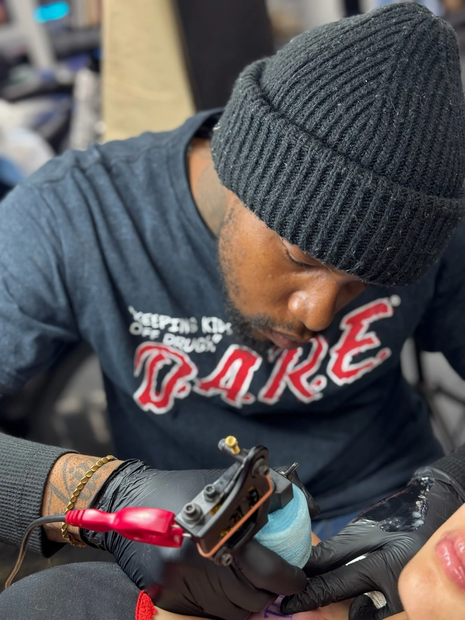 A person working on a project with a soldering iron, wearing black gloves and a gray beanie, with a tattoo on their forearm and a bracelet, inside a workshop or garage.