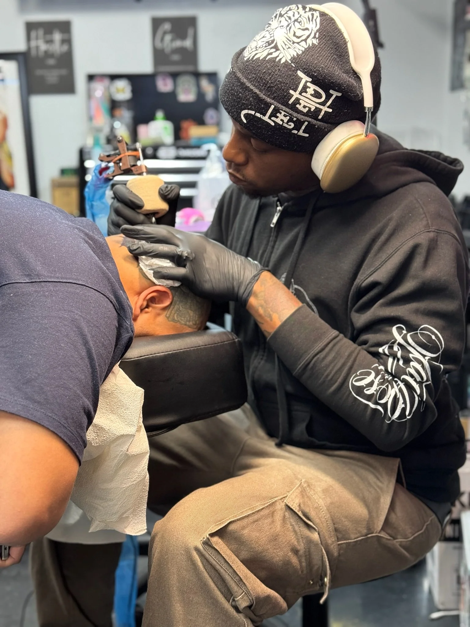 A person receives a tattoo on their upper arm while seated in a tattoo studio. The tattoo artist is wearing a black hoodie, beige cargo pants, gloves, and a beanie with a tiger graphic and the words 'T-shirt' and some Asian characters. The artist is 