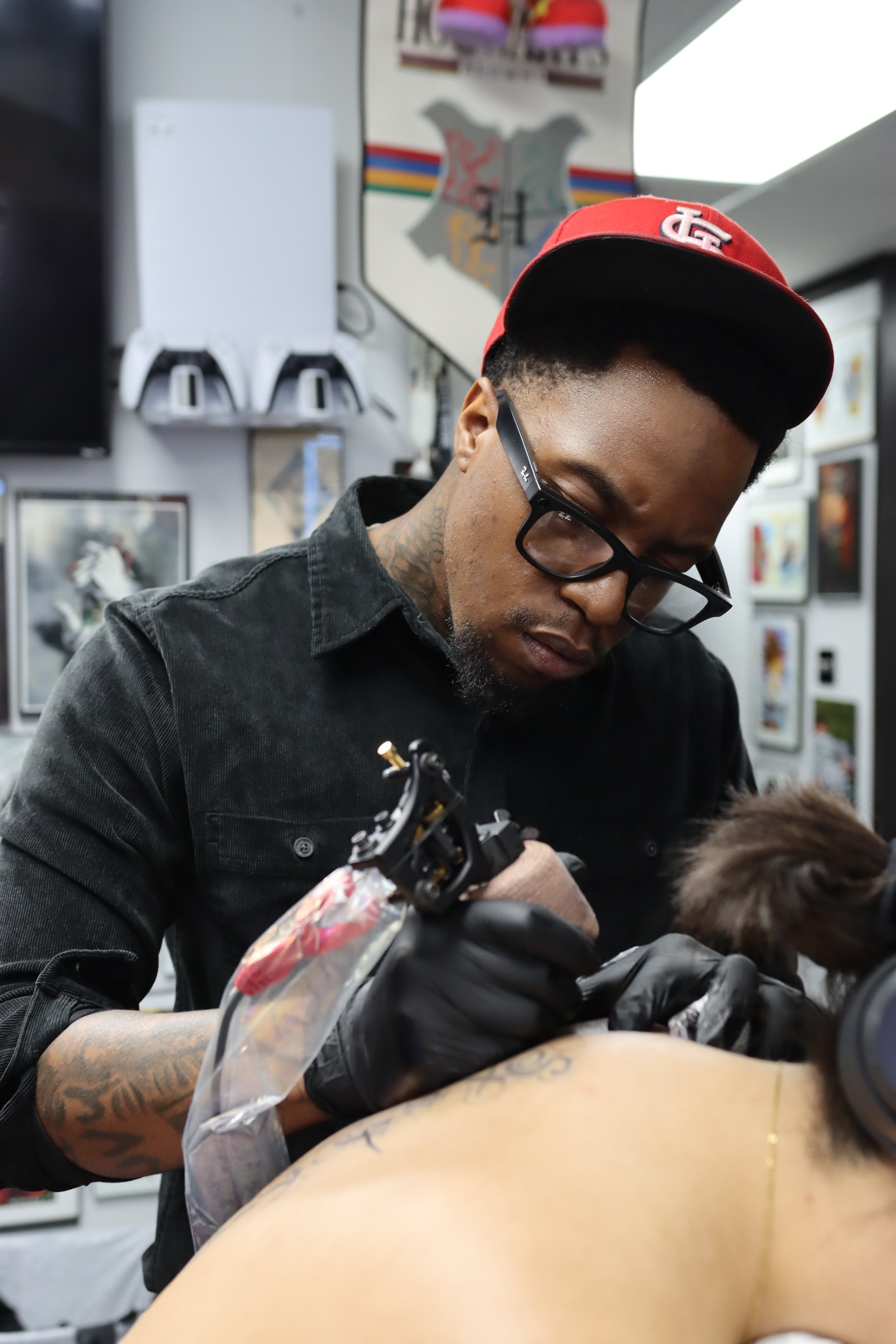 A tattoo artist wearing glasses and a red cap with white logo, working on a client's tattoo. The client is lying on a table with a plastic barrier and has multiple tattoos on their arm. The background has framed pictures and a decorative wall mirror.
