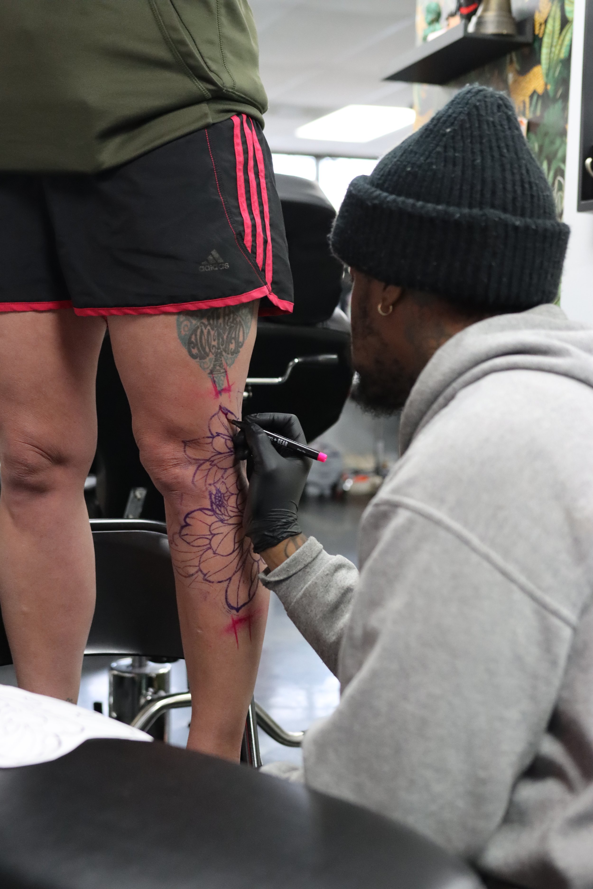 A person with a tattoo on their thigh is getting a tattoo on their leg by a tattoo artist wearing a gray hoodie and a black beanie in a tattoo studio.