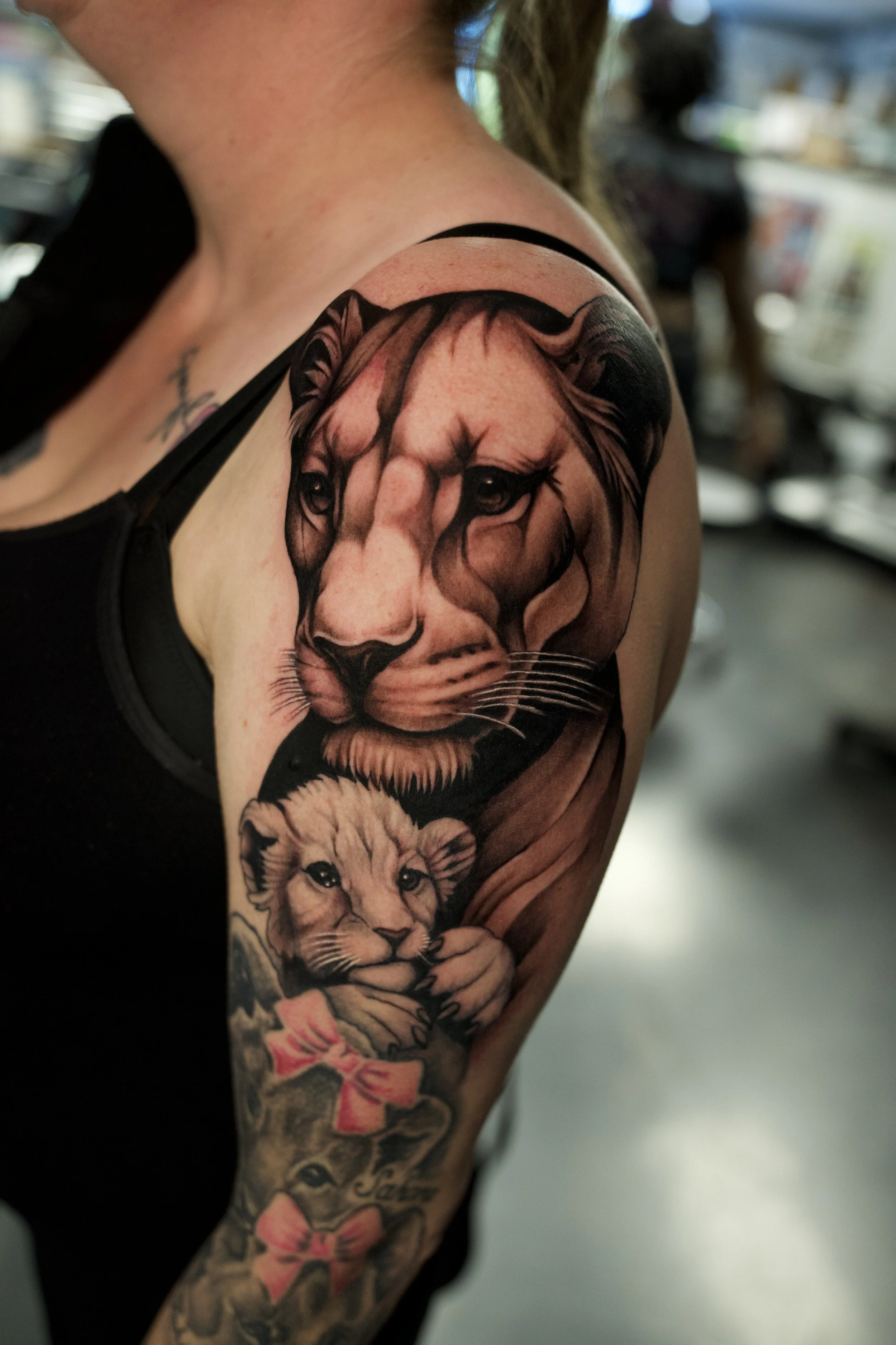 A close-up of a person's upper arm and shoulder, displaying a detailed, realistic tattoo of a lion and a lioness on the arm.
