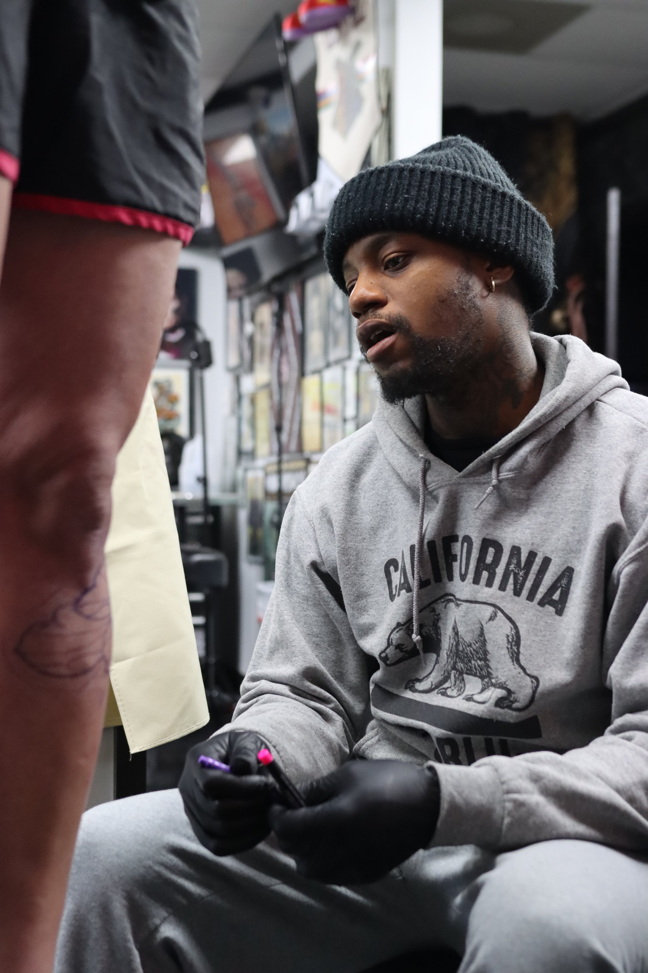 A man with a beard, wearing a gray hoodie with a bear graphic and the word 'California,' and a black knit beanie, is sitting and looking at a person who is standing nearby. The man is holding a purple marker and appears to be signing or drawing somet