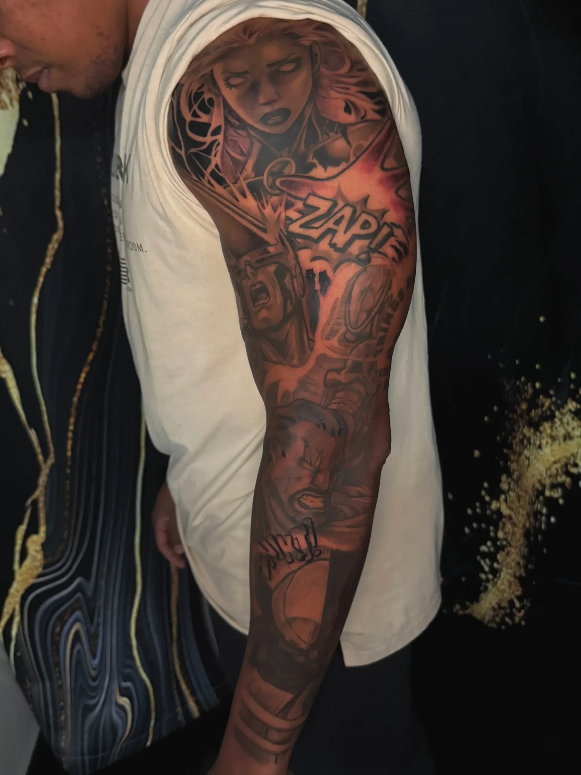 A person with a large colorful tattoo sleeve on their arm, wearing a white tank top, and holding a drink, possibly a beer, with a patterned background.