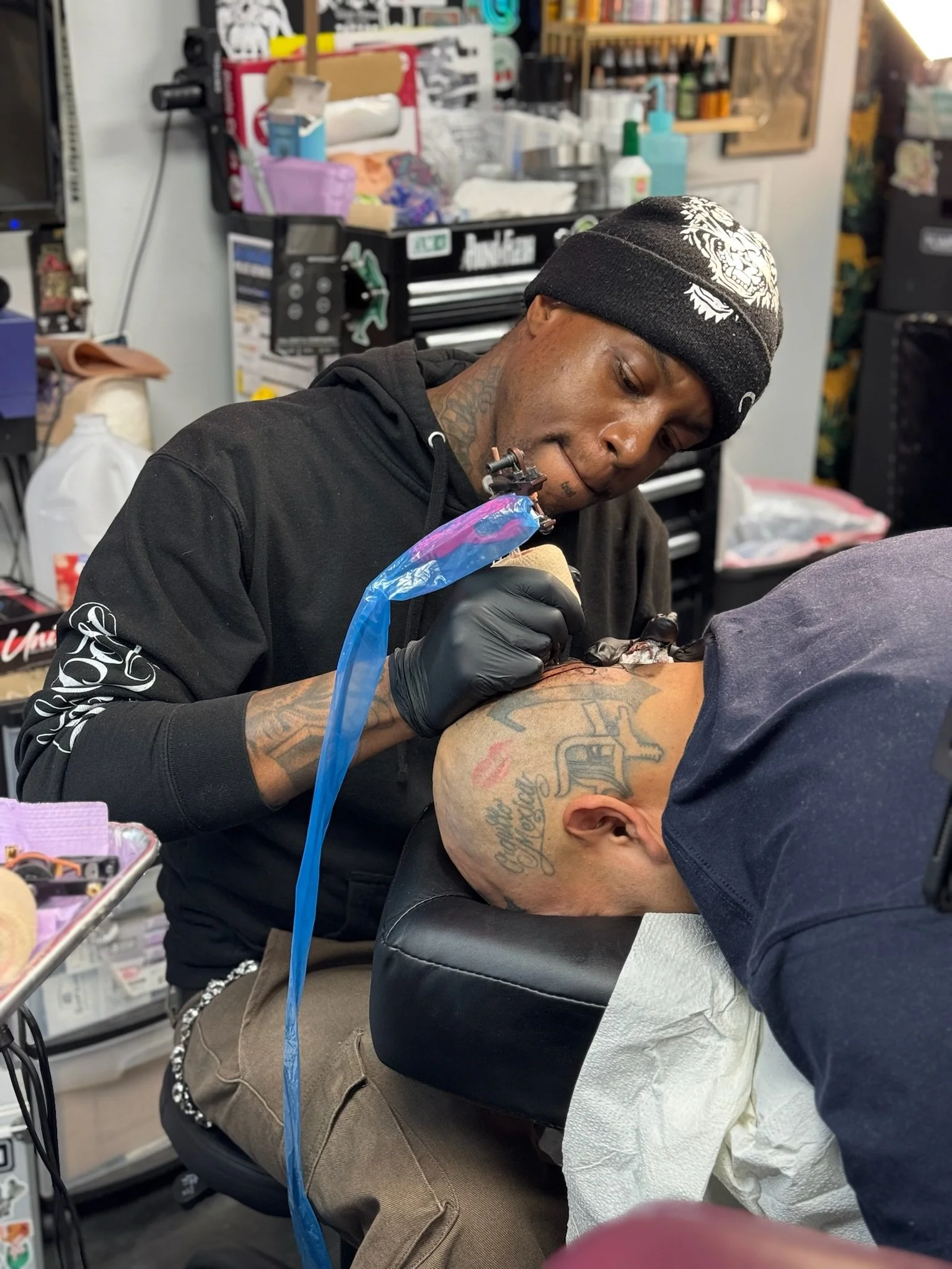 A tattoo artist wearing a black beanie and black gloves tattoos a person's head in a tattoo shop, with various supplies and equipment in the background.