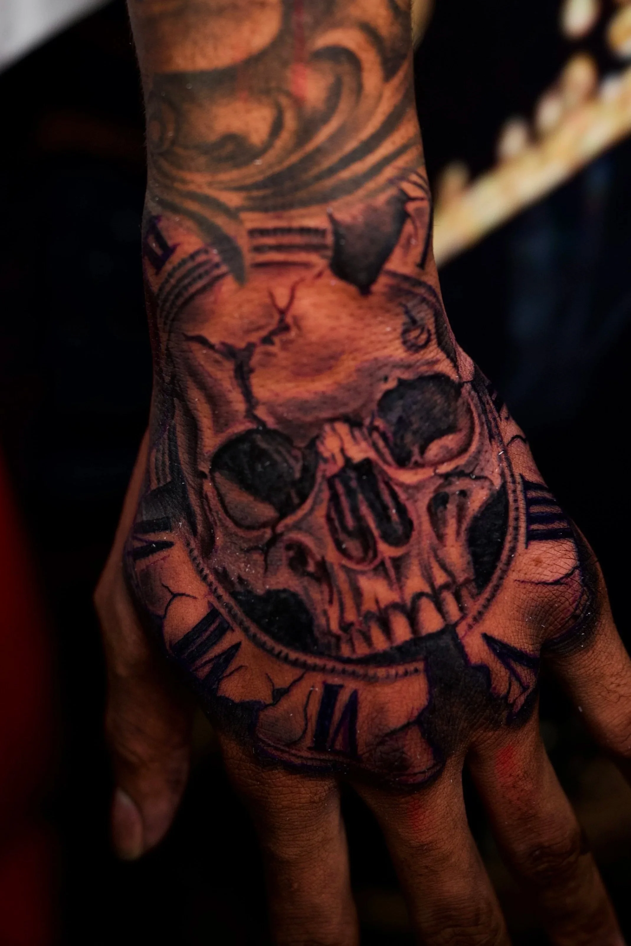 Close-up of a hand with a detailed tattoo covering the entire hand and wrist. The tattoo features various designs, including a skull, wings, and a shield with text. The skin appears slightly distressed or wrinkled.