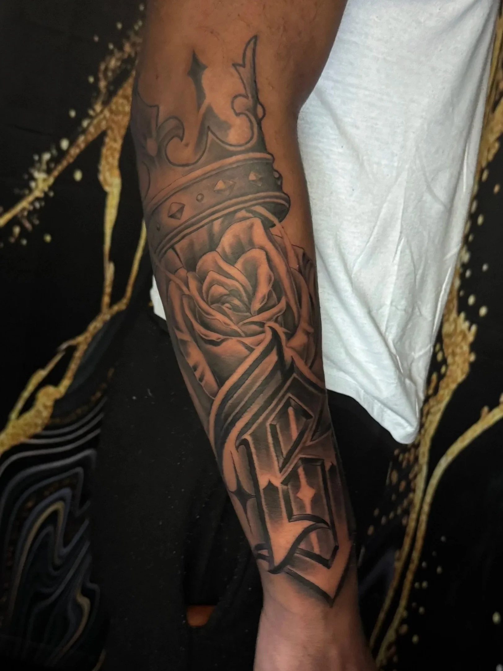 A person's arm with a detailed black and gray tattoo sleeve featuring a crown, a rose, and stars.