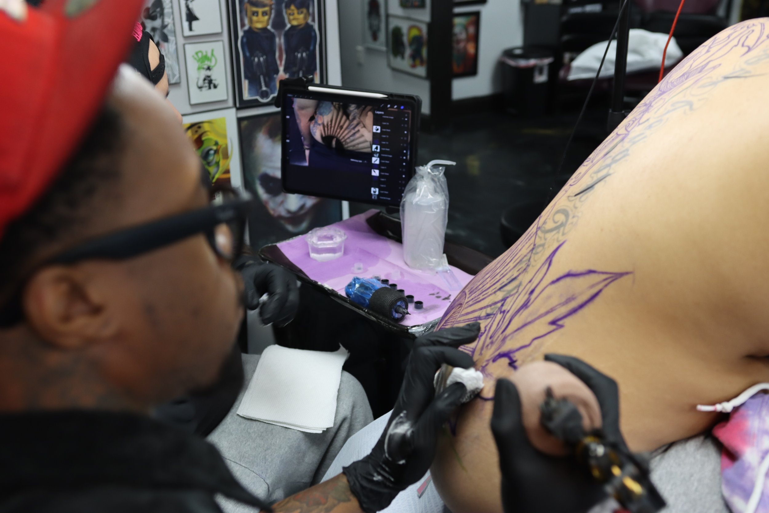 Tattoo artist working on a person's leg in an art studio, with framed artwork on the wall in the background.