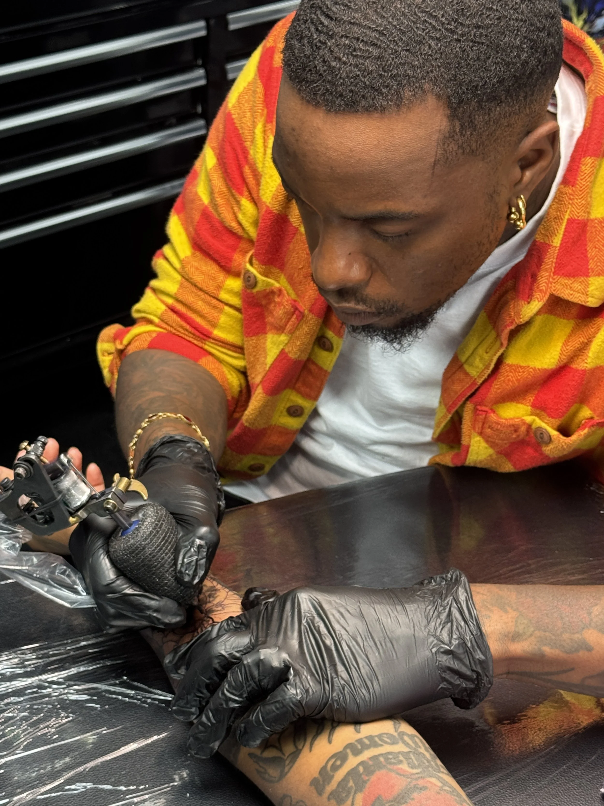 A tattooed person with short hair and earrings getting a tattoo on their leg in a tattoo studio while wearing a yellow and red checkered flannel shirt. The tattoo artist wears black gloves and is working with a tattoo machine.