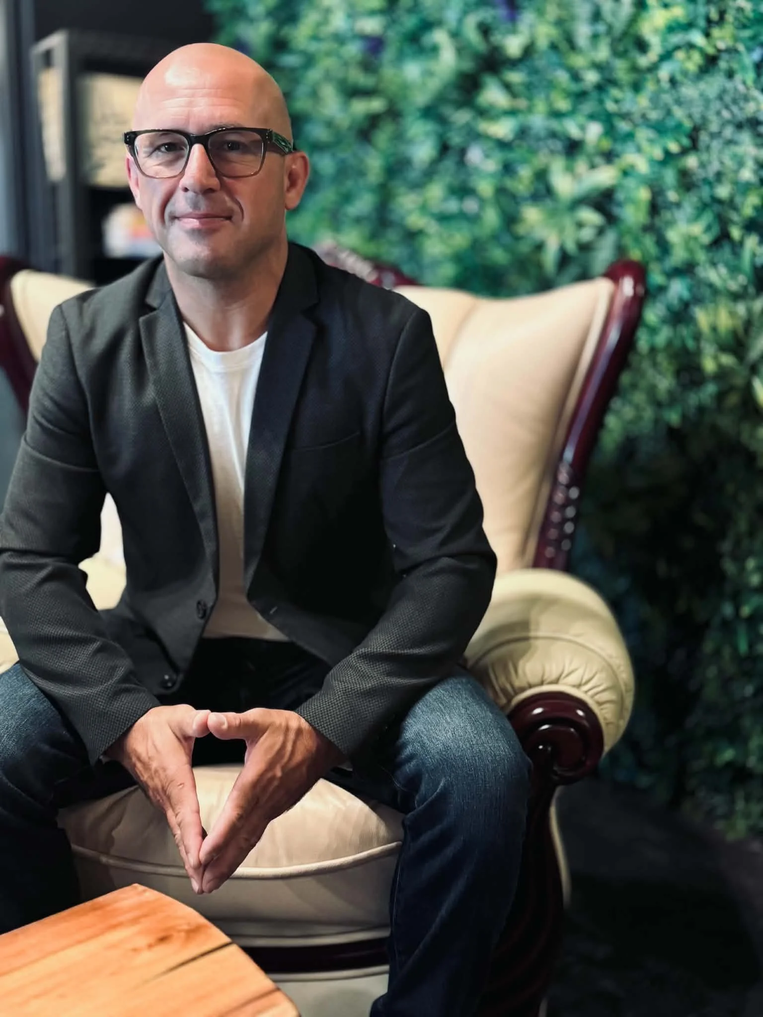 A bald man with glasses sitting on a beige sofa with a green leafy mural in the background. The man's hands are clasped together, resting on his lap, and he is wearing a black blazer and white t-shirt.