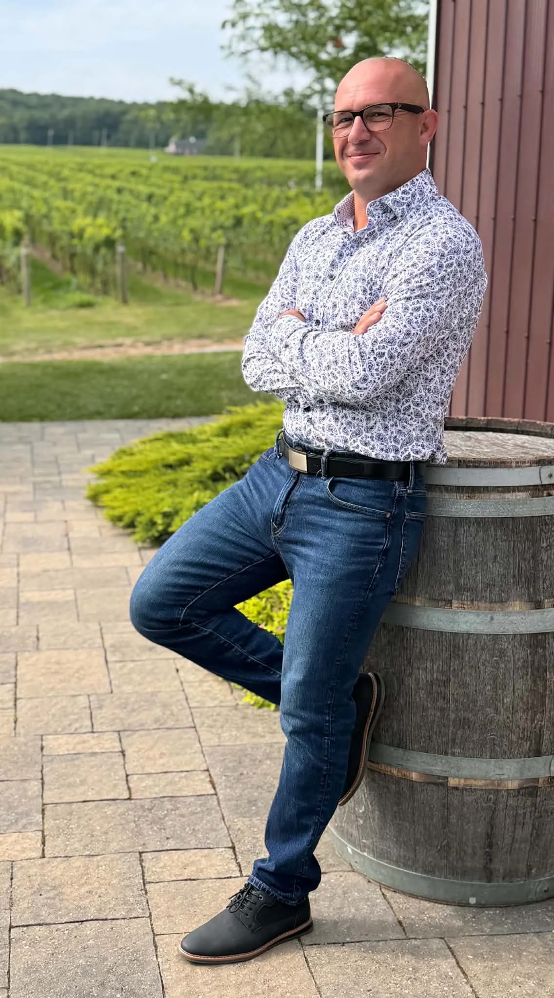 A man with glasses, wearing a patterned shirt and jeans, stands outdoors leaning against a large wooden barrel with crossed arms, in front of a vineyard and trees.