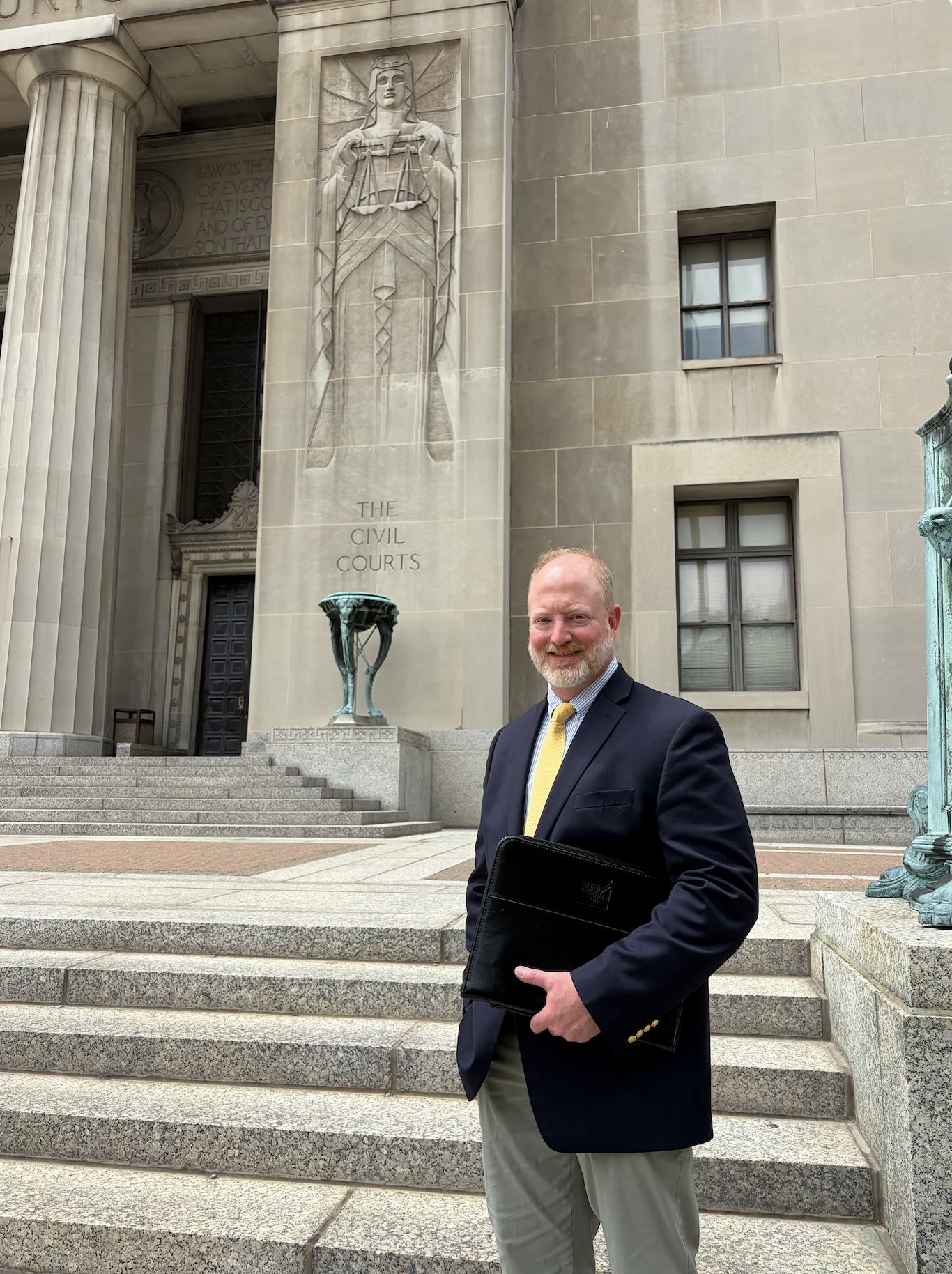 A man in a business suit holding a black folder standing in front of the Civil Courts building with large stone steps, columns, statues, and engraved text on the building.