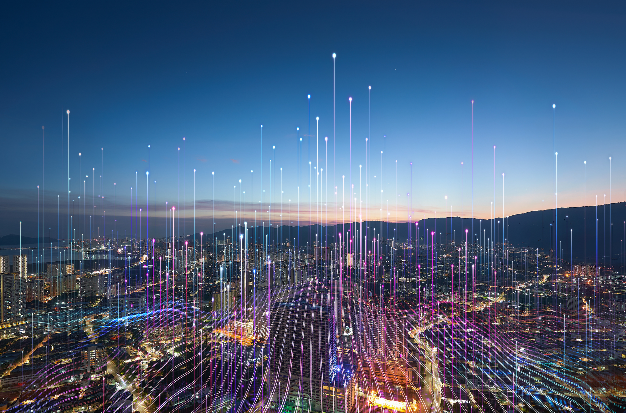 City skyline at dusk with colorful light trails resembling digital data or fiber optic lines streaming upward, creating a futuristic visual effect.