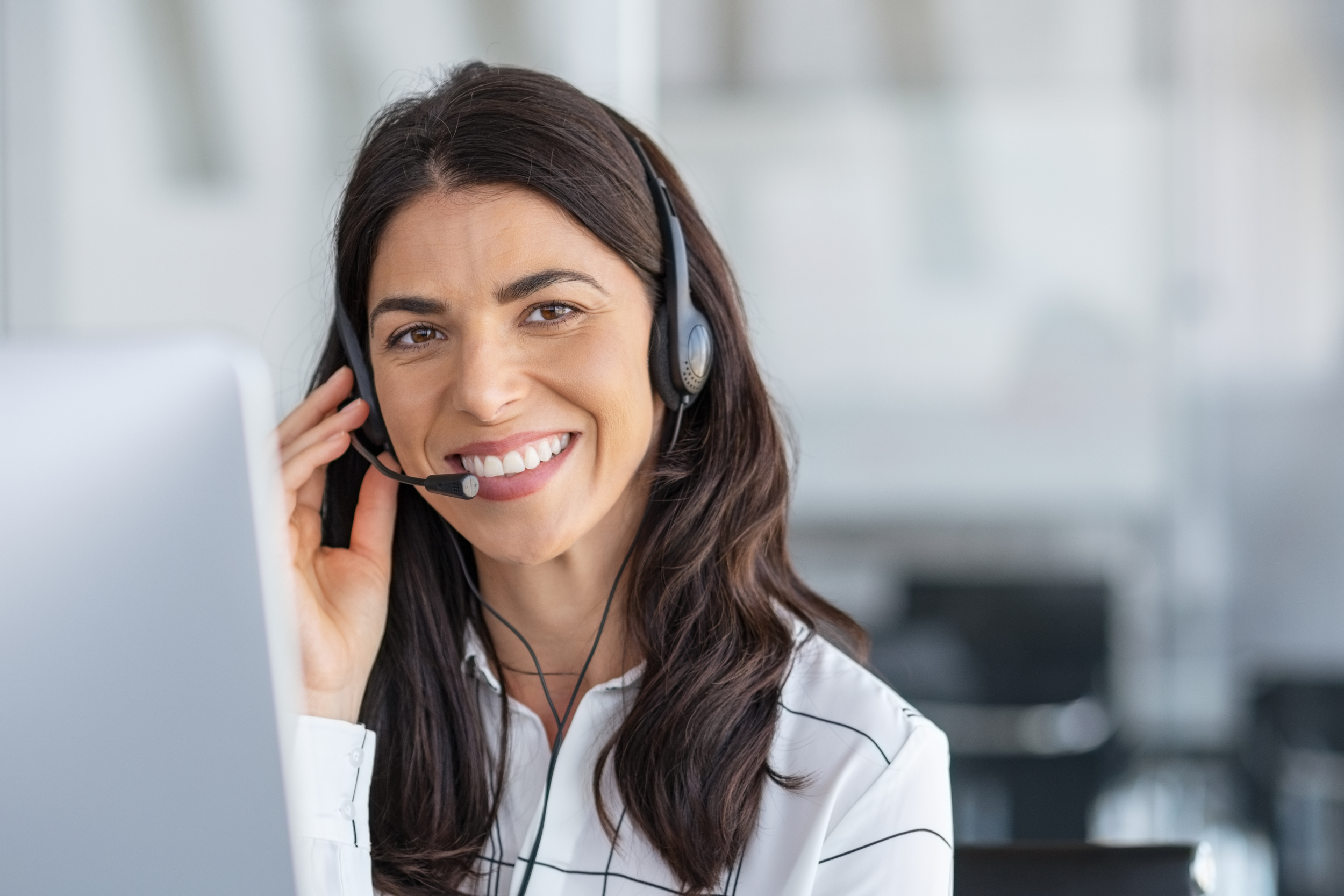 A woman with long dark hair wearing a white blouse and headset, smiling at a computer screen in an office setting.