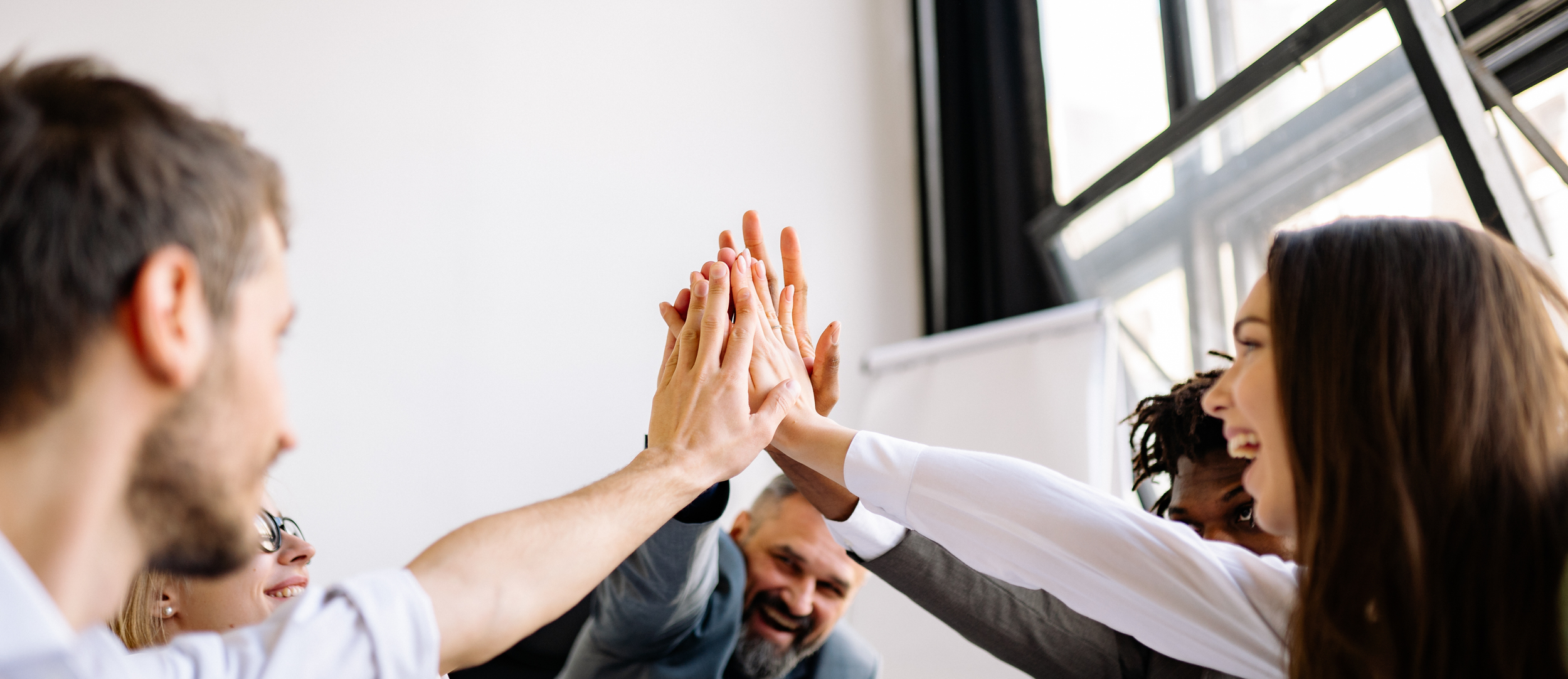 Group of diverse people giving high five in a bright room with large windows.