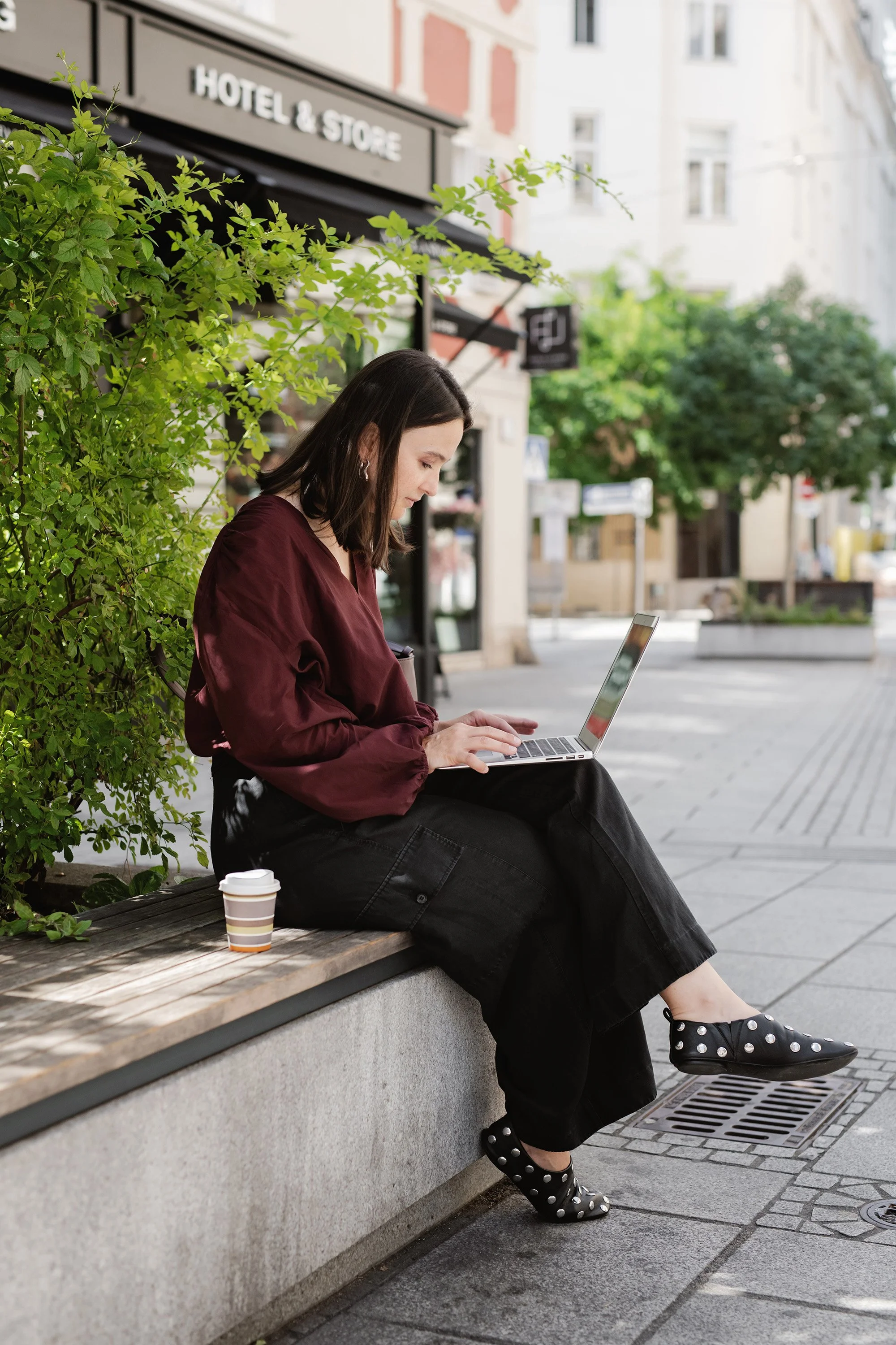 A young woman with dark hair sitting on a wooden bench outdoors, wearing a maroon jacket, black pants, and polka dot shoes, working on a laptop. There is a disposable cup on the bench next to her. Green bushes and trees are in the background, along with buildings and a sidewalk.