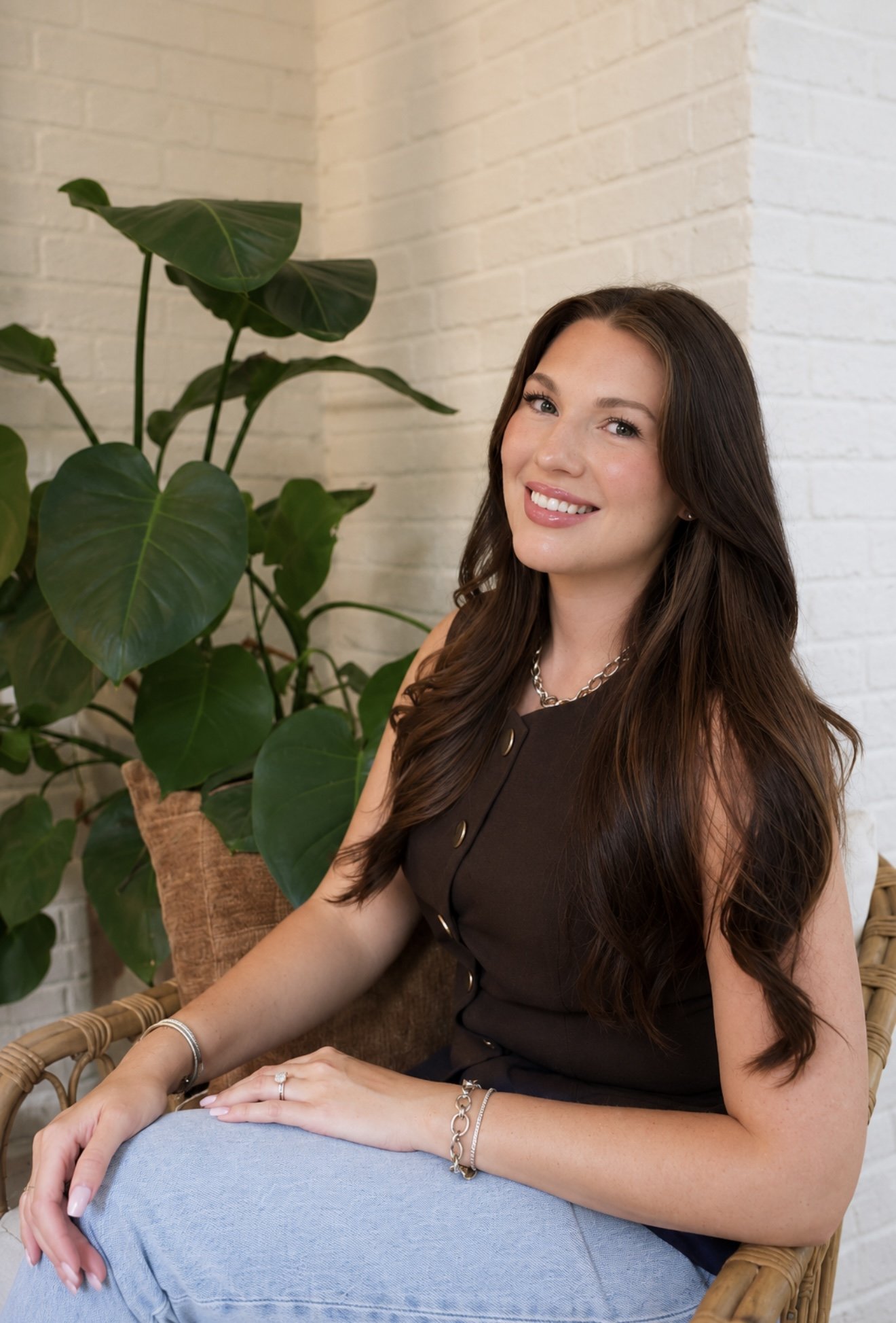 A woman with long, wavy brown hair sitting in a wicker chair with green plants and a white brick wall in the background.