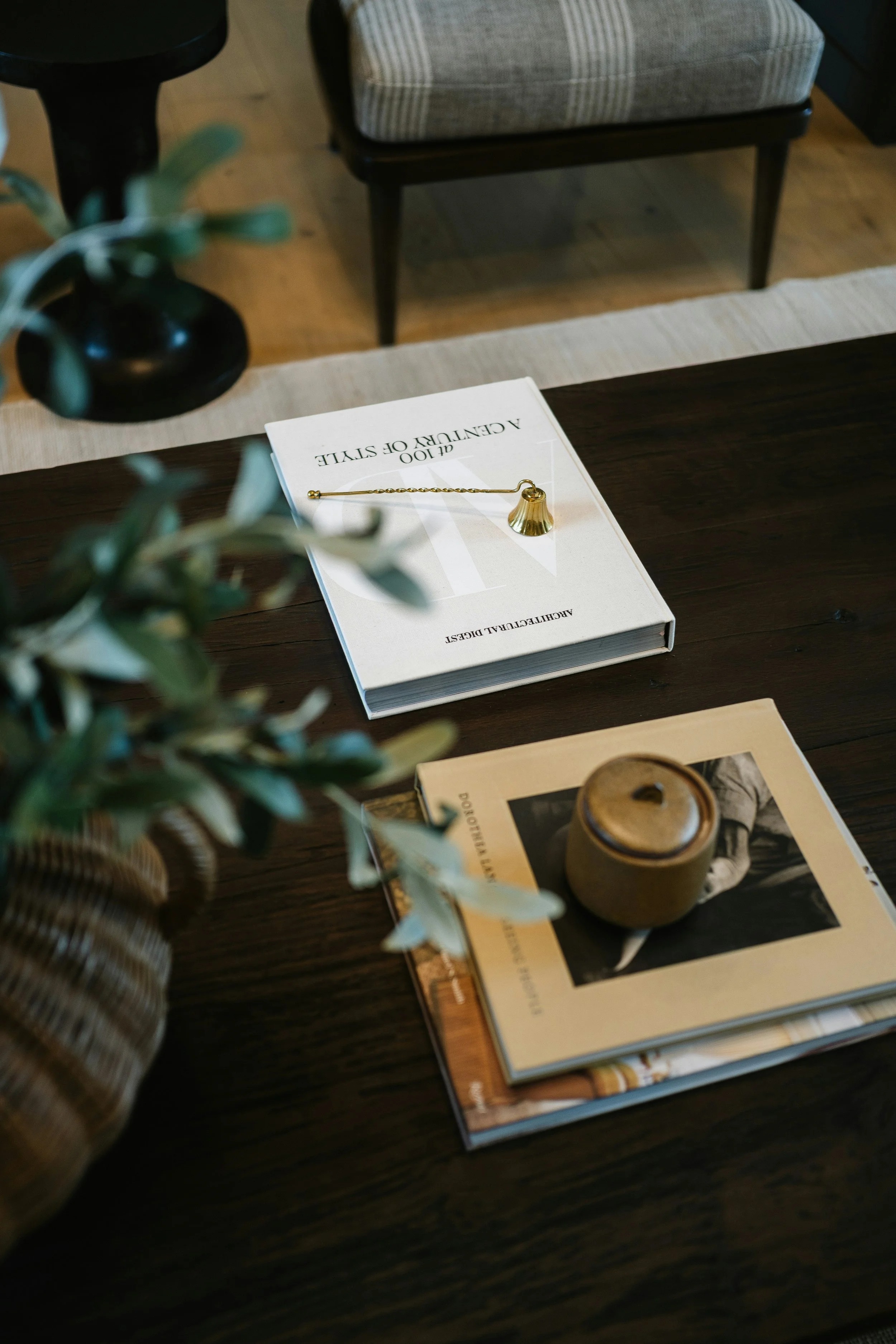 A wooden table with a white book titled 'A Century of Style' on top of magazines and a small ceramic container. In the background, a chair with striped upholstery and wooden legs can be seen, along with a black table or object. There is also a blurred green plant in the foreground.
