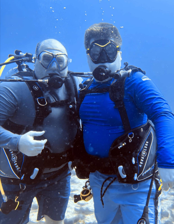 Two scuba divers underwater, wearing wetsuits, masks, and snorkels, posing for a photo while giving a thumbs-up.