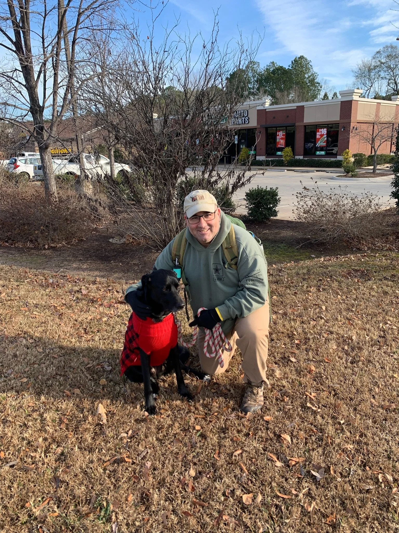 A man in outdoor clothing kneeling on grass with a black dog wearing a red sweater. In the background, there are parking lot and storefronts with signs, and leafless trees, indicating a late fall or winter setting.