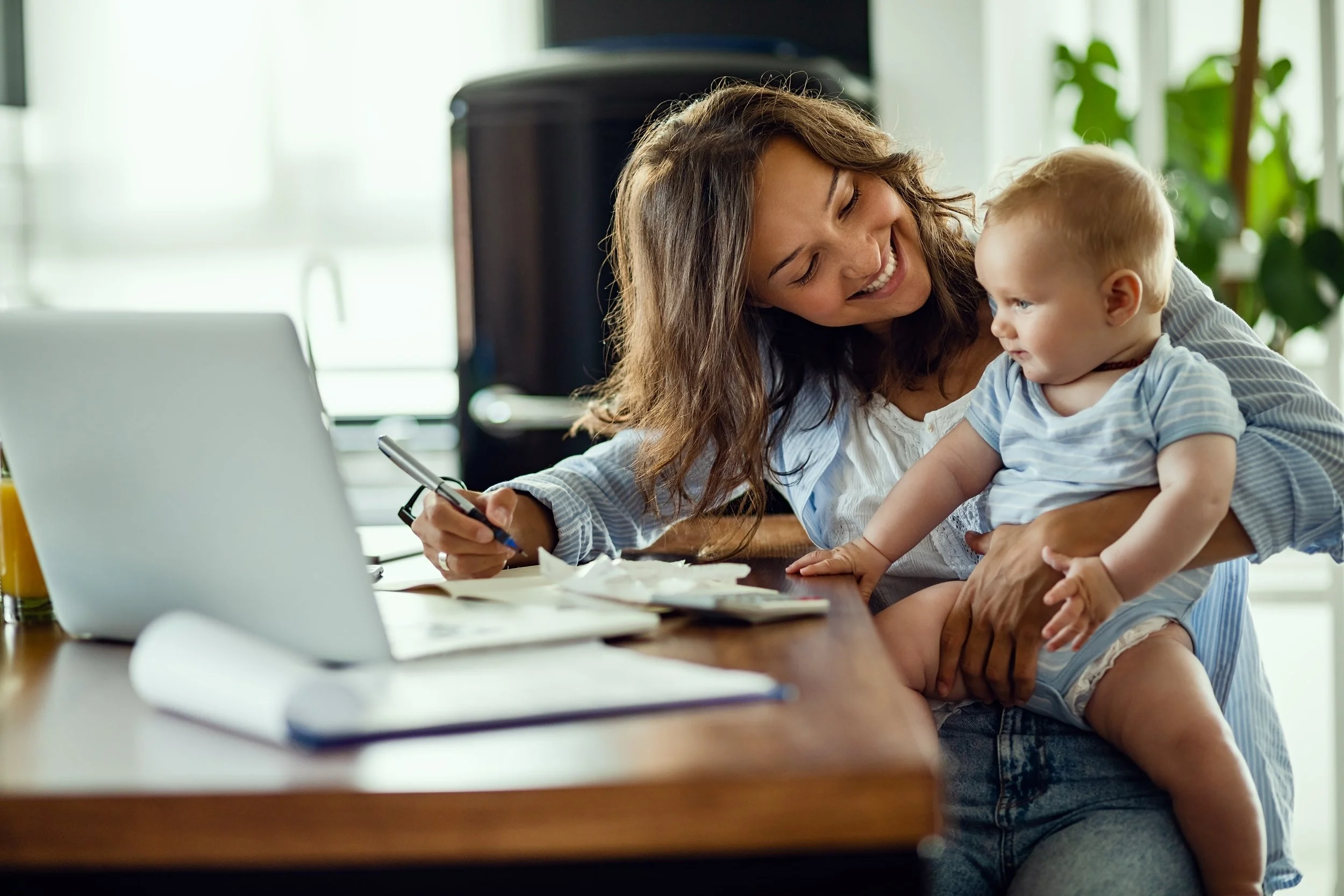 A woman with long brown hair smiling at a young child with blonde hair, who is looking at her. They are at a wooden table with papers, a pen, and laptops, in a bright room with green plants.