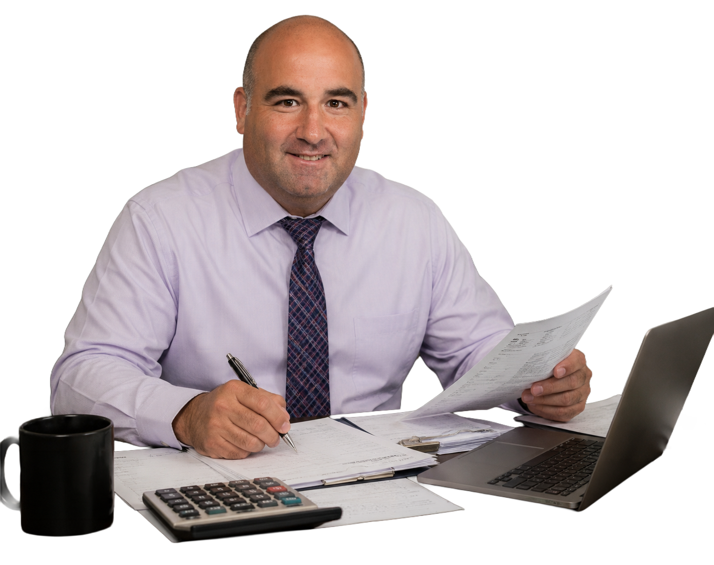 A man sitting at a desk with papers, a calculator, a laptop, and a coffee mug, holding a document and a pen, smiling at the camera.