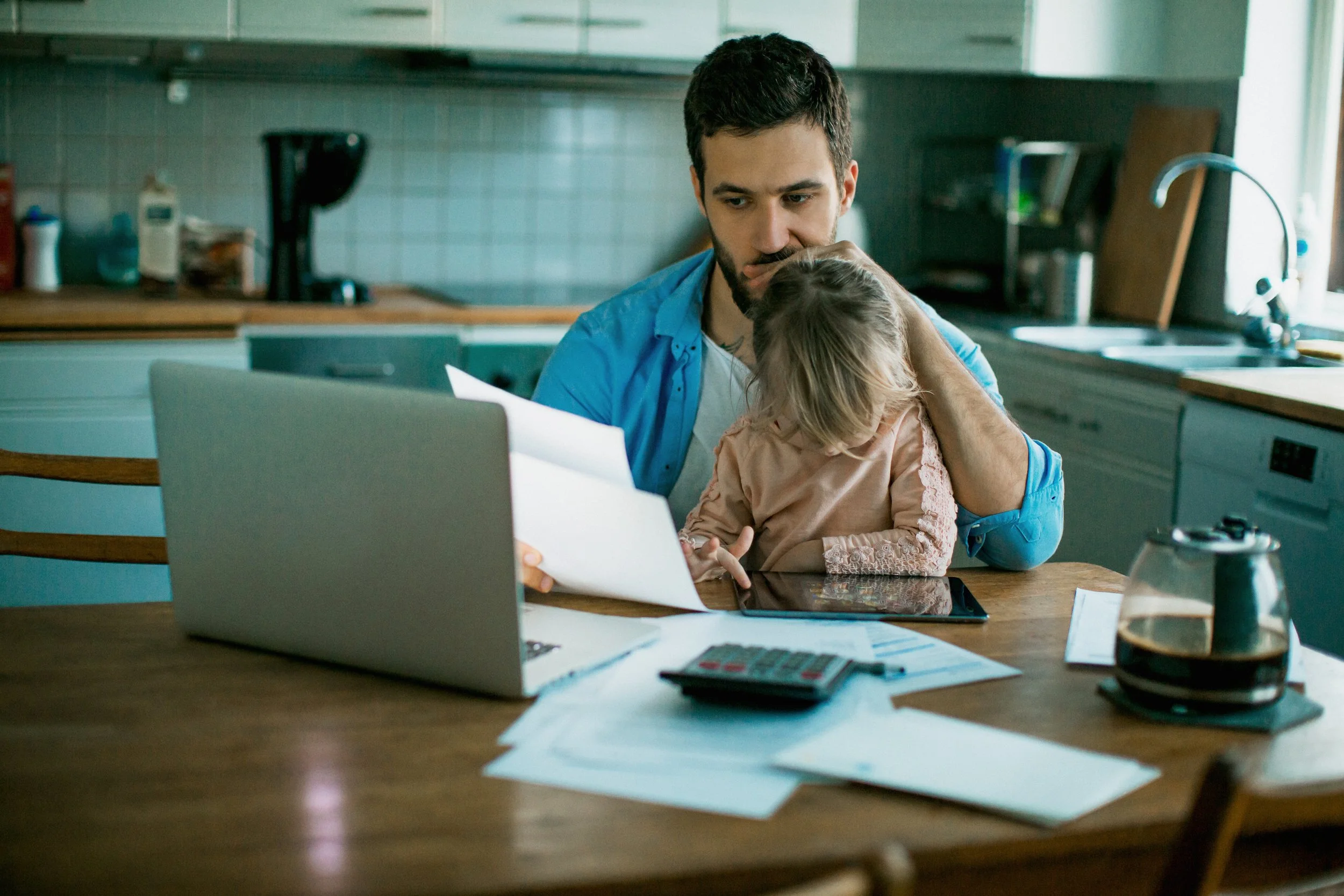 A man and a young girl sitting at a cluttered kitchen table looking at a laptop screen, with papers, calculator, and various items scattered on the table.