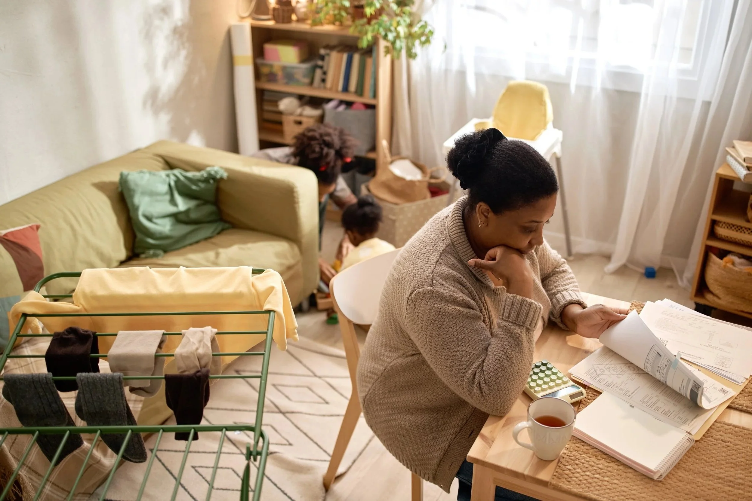A woman sitting at a table reading papers, with a cup of tea in front of her. In the background, two children playing near a shelf with books and storage boxes. A drying rack with socks and underwear is in the foreground, and a sofa with a green blanket is on the left side of the room. The room has natural light coming through sheer curtains.