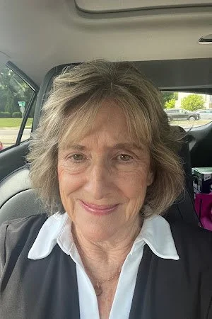 A woman with short, wavy blonde hair sitting in a car, smiling at the camera. She is wearing a white collared shirt and a dark vest or jacket.