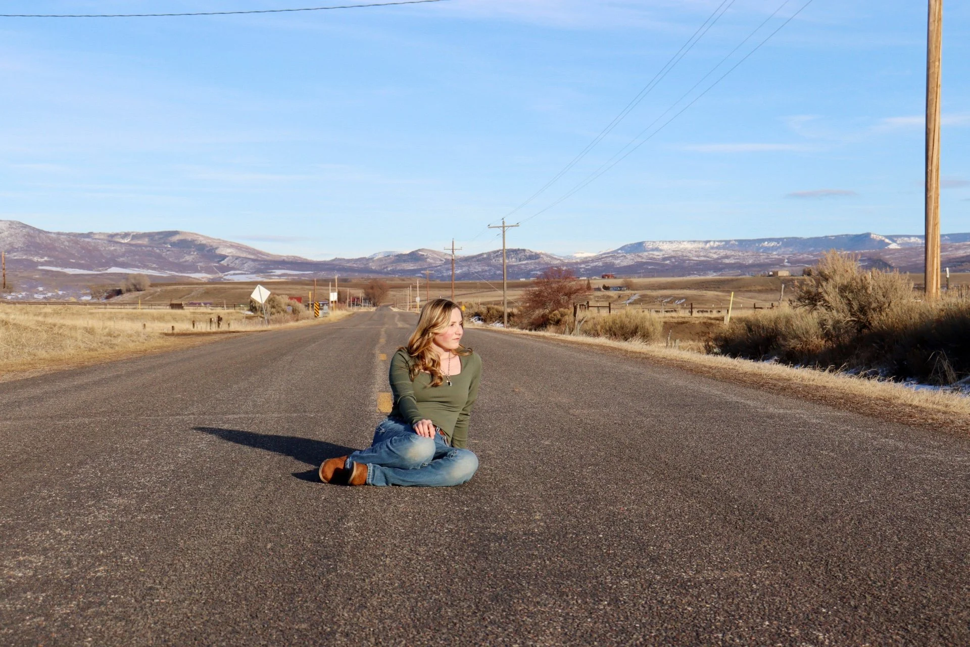 Western Edge Studios. A woman with blonde hair sitting cross-legged on an empty rural road in a mountainous area with blue skies, dry grass, and power lines in the background.
