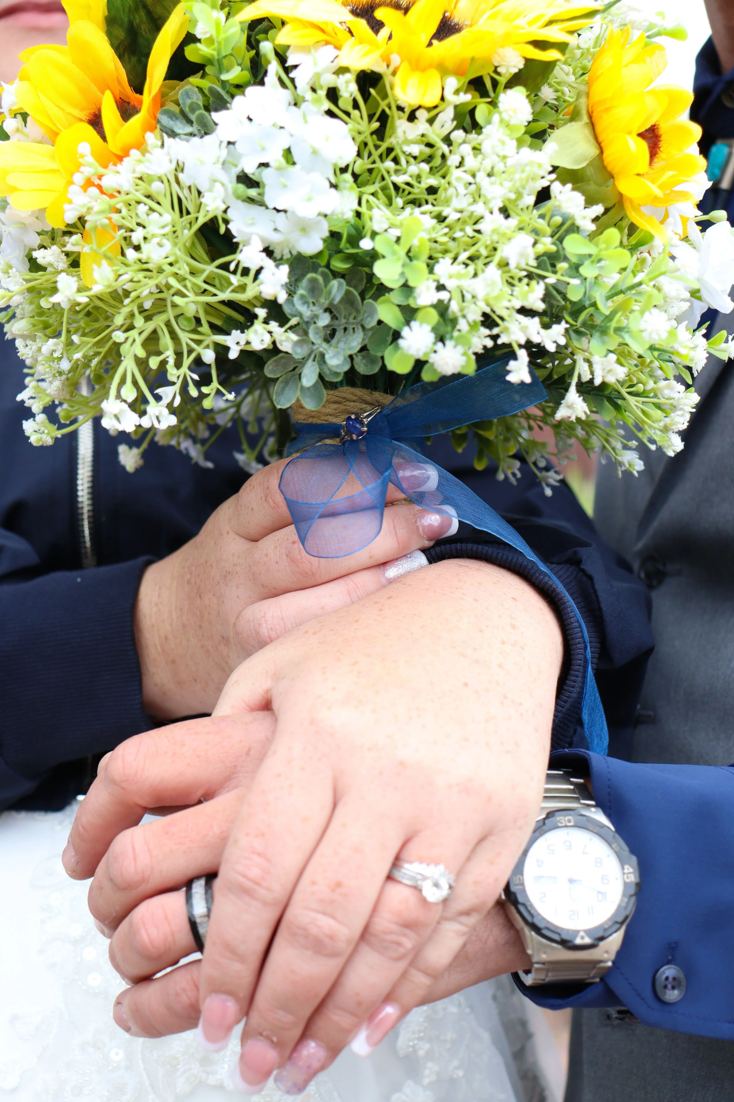 Western Edge Studios. Close-up of a wedding bouquet with yellow sunflowers and white flowers, held by a bride and groom holding hands, with rings visible, against a blurred background of people.