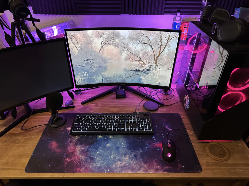 Western Edge Studios. A computer desk setup with two monitors, a keyboard, a mouse, a microphone, and a gaming PC with pink LED lights. The desk has a galaxy-themed mouse pad, and the background features purple acoustic foam panels and LED lighting.