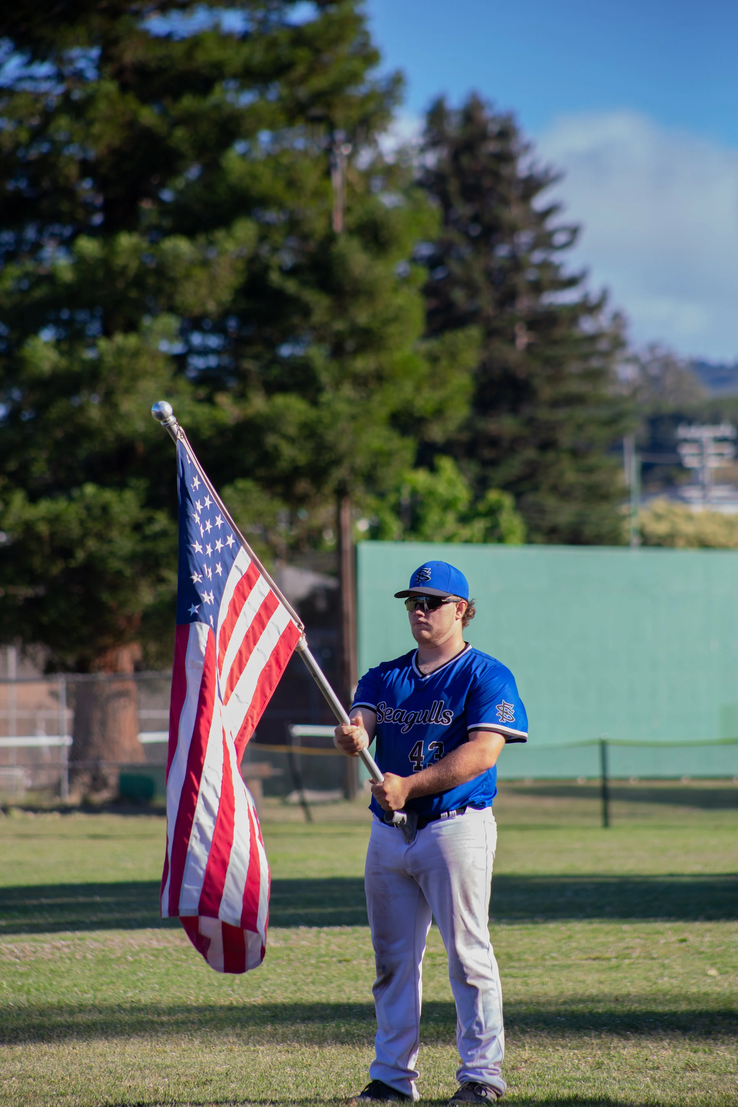 A baseball player wearing a blue uniform with the number 42 on it, holding an American flag. The player has a blue cap and sunglasses, standing on a grassy field with trees and a tennis court in the background.