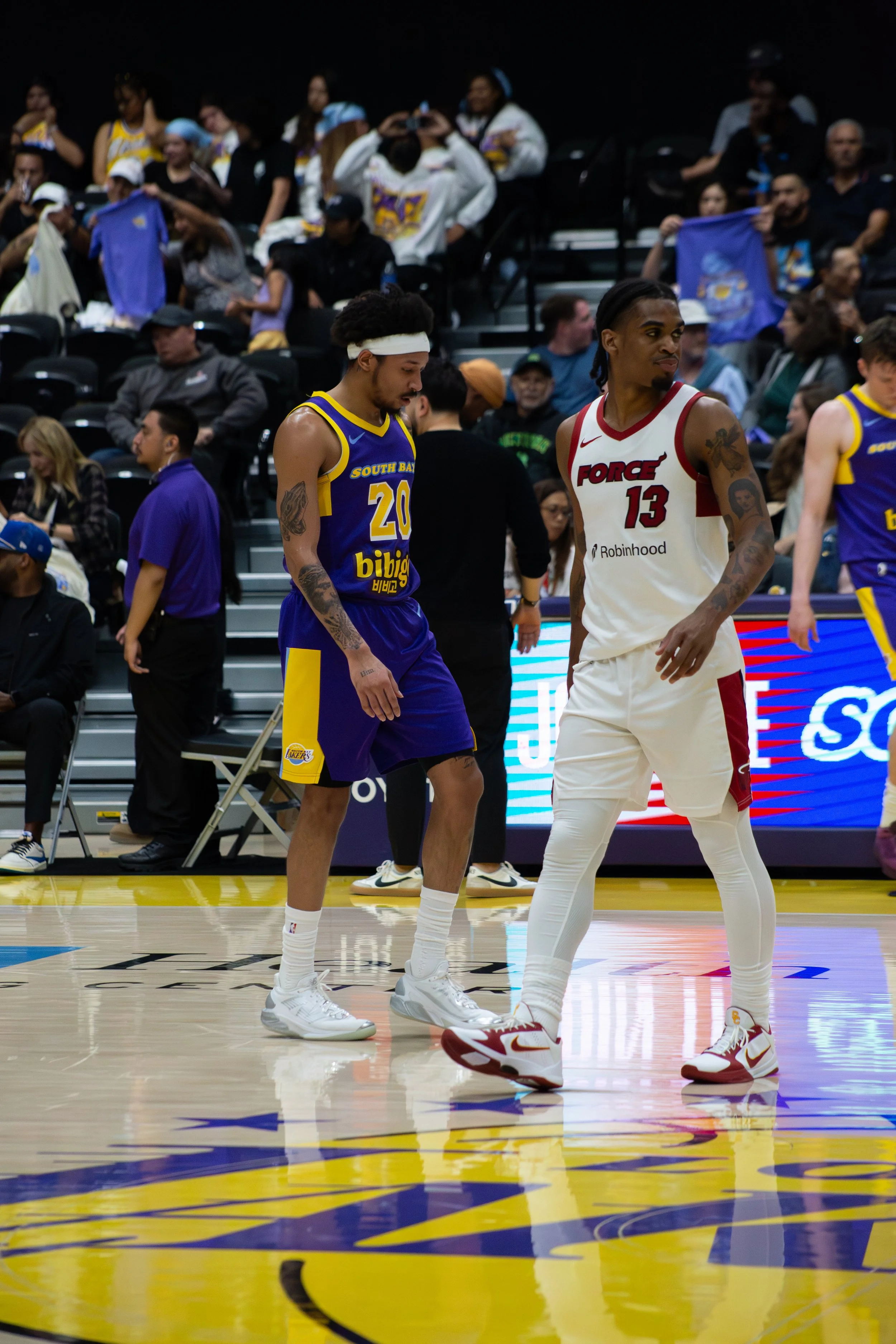 Two basketball players standing on the court, one in a yellow and purple uniform and the other in a white and red uniform, with a crowd watching in the background.