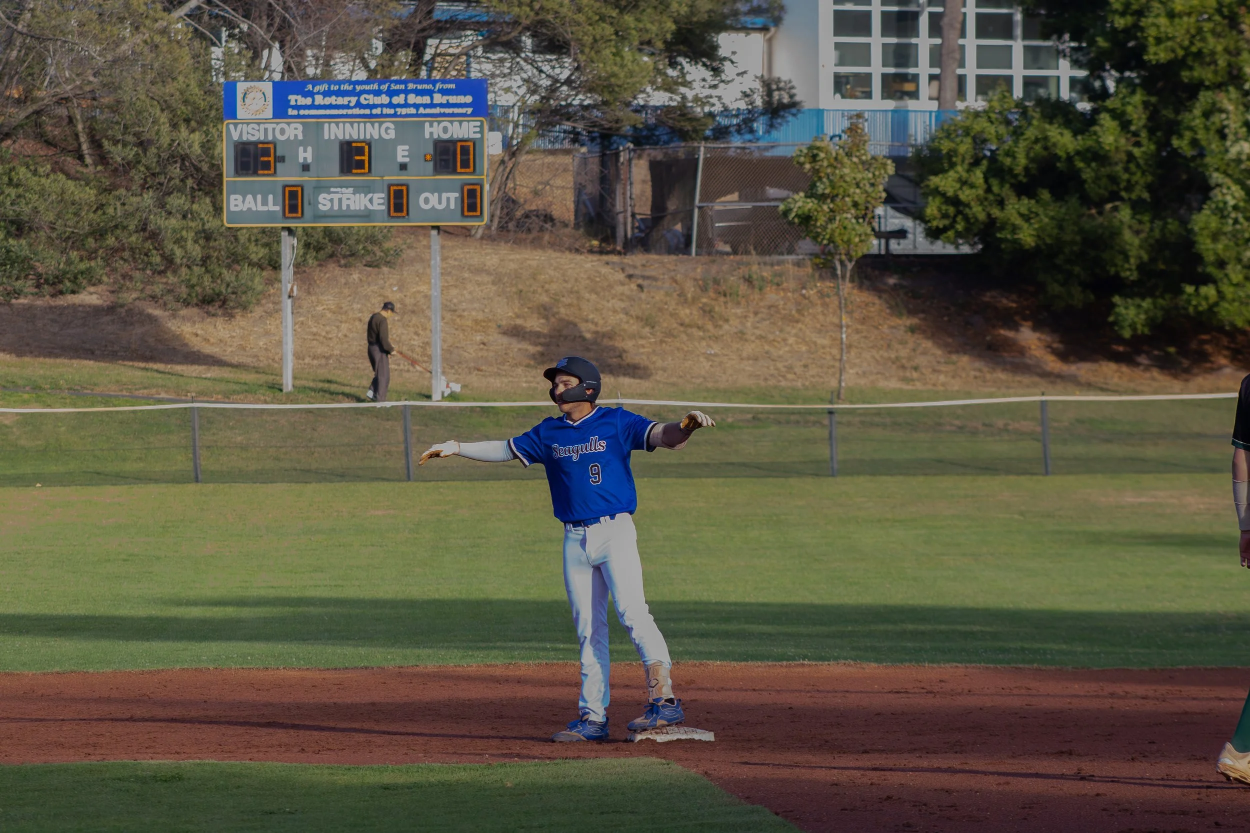 A baseball player wearing a blue uniform with the number 9 is on base, standing on a base on the field, with a baseball diamond in the foreground. In the background, a scoreboard shows the score as 3-3 with 3 outs, and a few people are on the infield