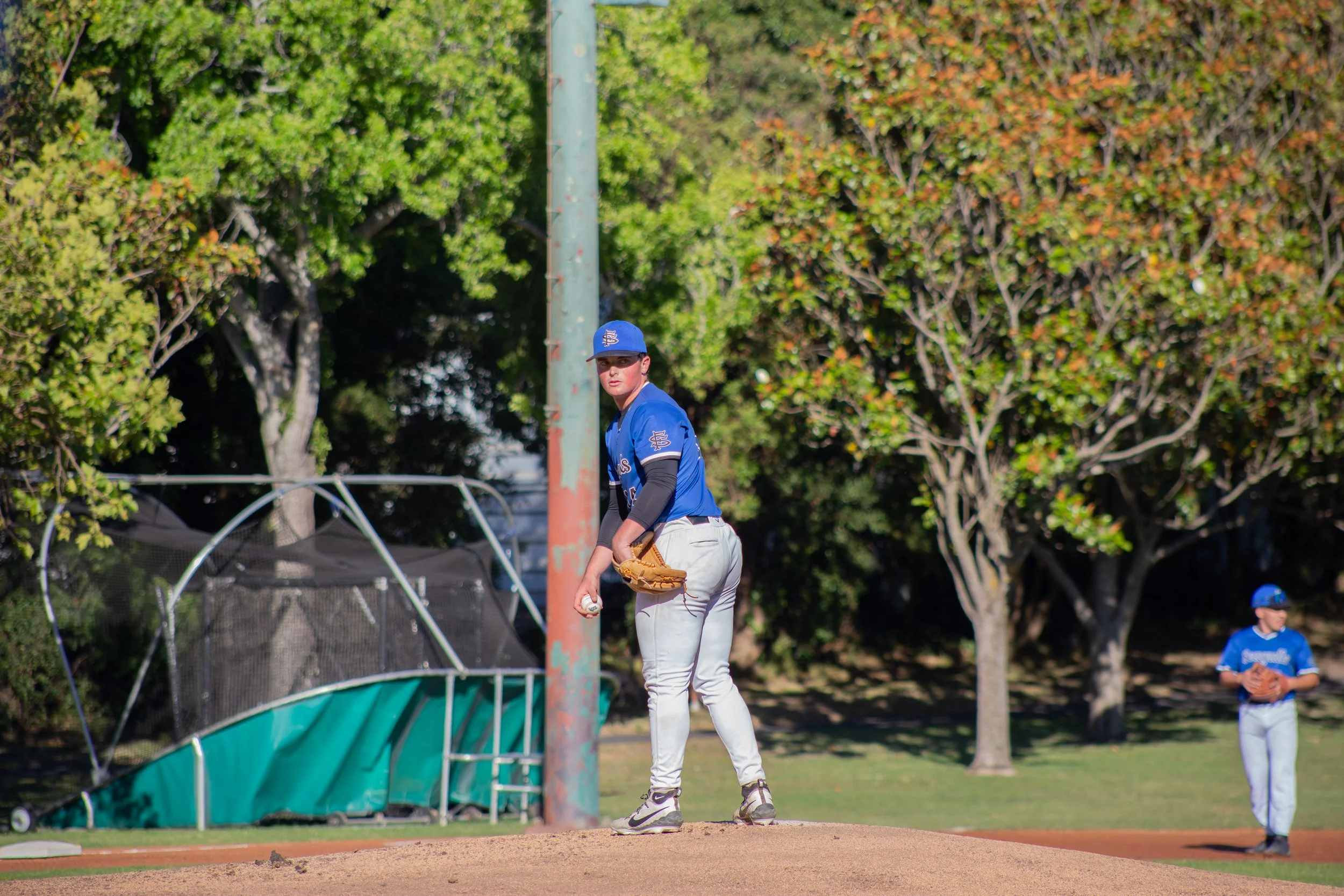 A young baseball player in a blue uniform and cap stands on a baseball field, holding a baseball in one hand and a glove on the other, near a tall pole with a green fence and trees in the background.