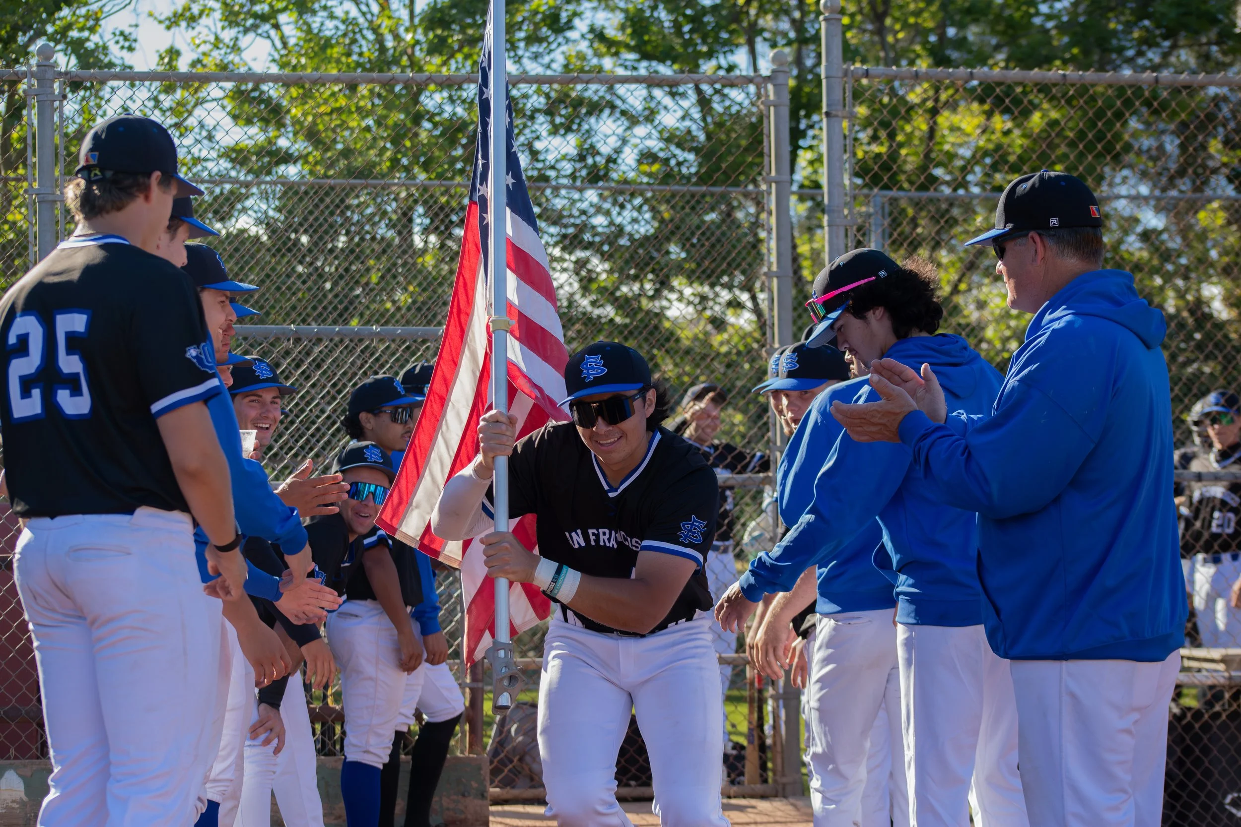 Youth baseball team gathered around an American flag, with players dressed in blue and white uniforms and black hats, during a ceremony at a baseball field on a sunny day.