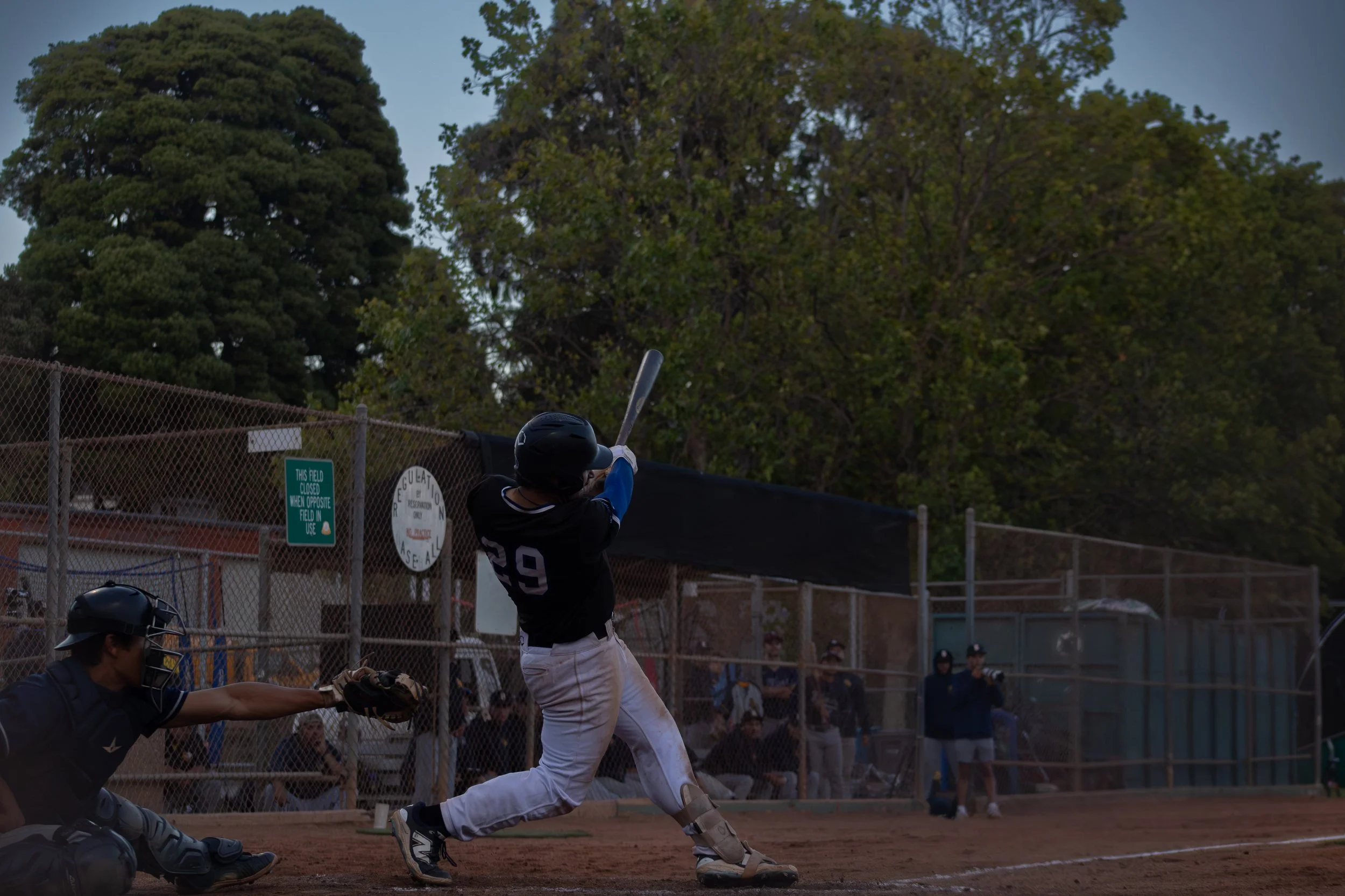 A baseball batter in black jersey and white pants swings at a pitched ball during a game at a baseball field, with a catcher and umpire behind him and green trees in the background.