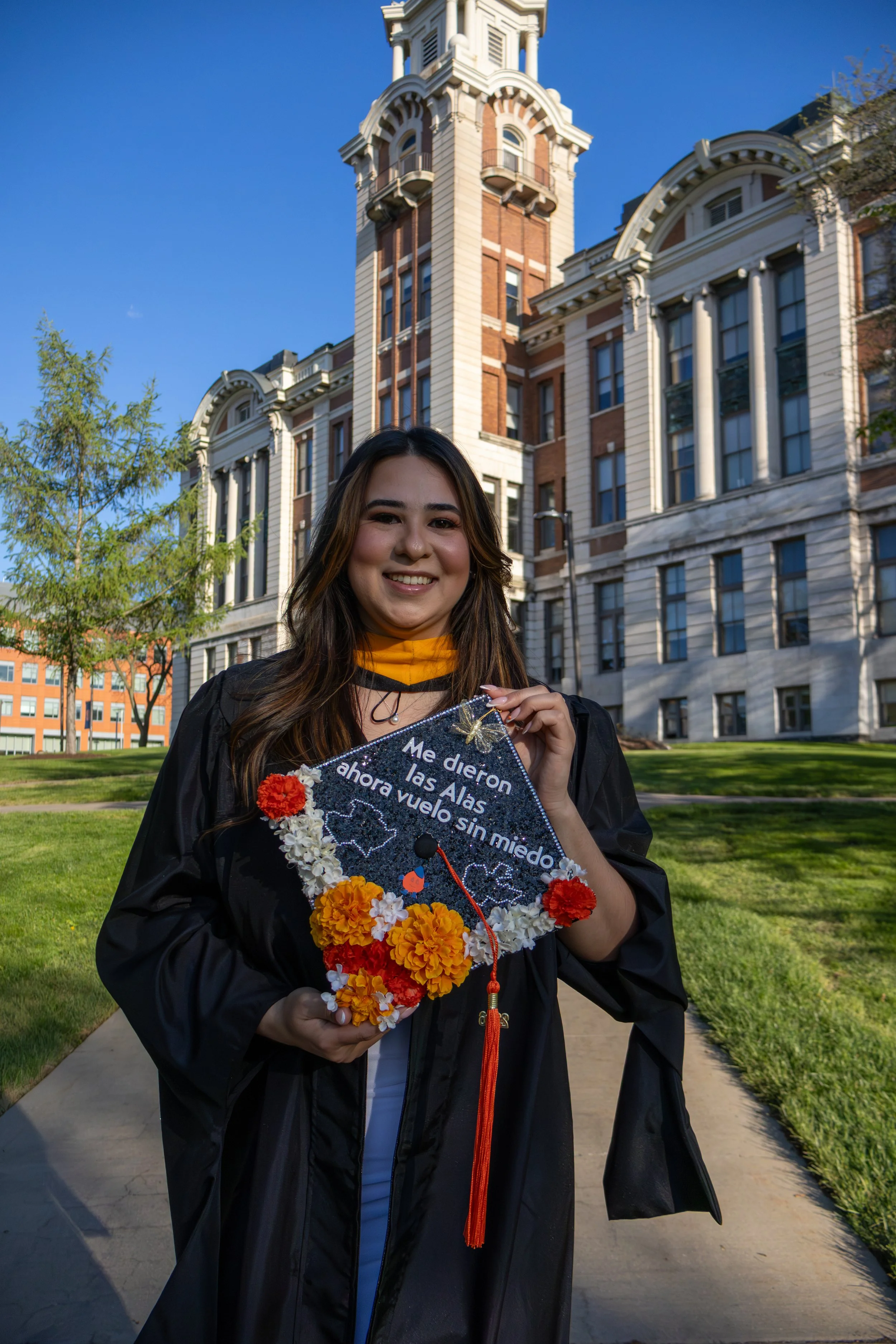 A young woman smiling in her graduation gown and cap, holding a decorated stole that says "Me dieron las Alas ahora vuelvo sin miedo" with a map of Mexico and colorful flowers. She stands on a pathway in front of a historic building with a clock towe