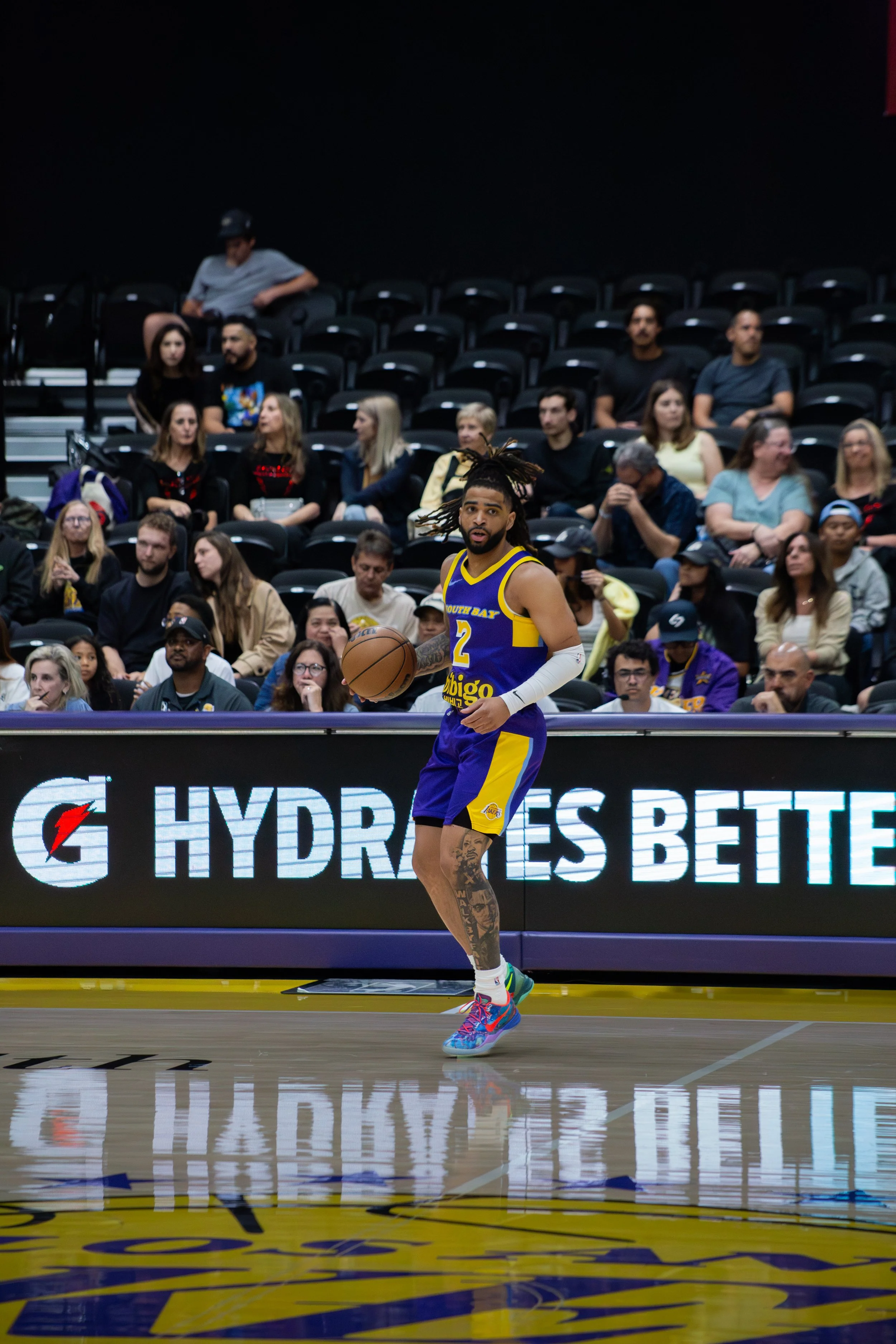 A basketball player with long hair and tattoos on his leg, wearing a blue and yellow jersey with the number 2, dribbling a basketball on a basketball court with spectators in the background.