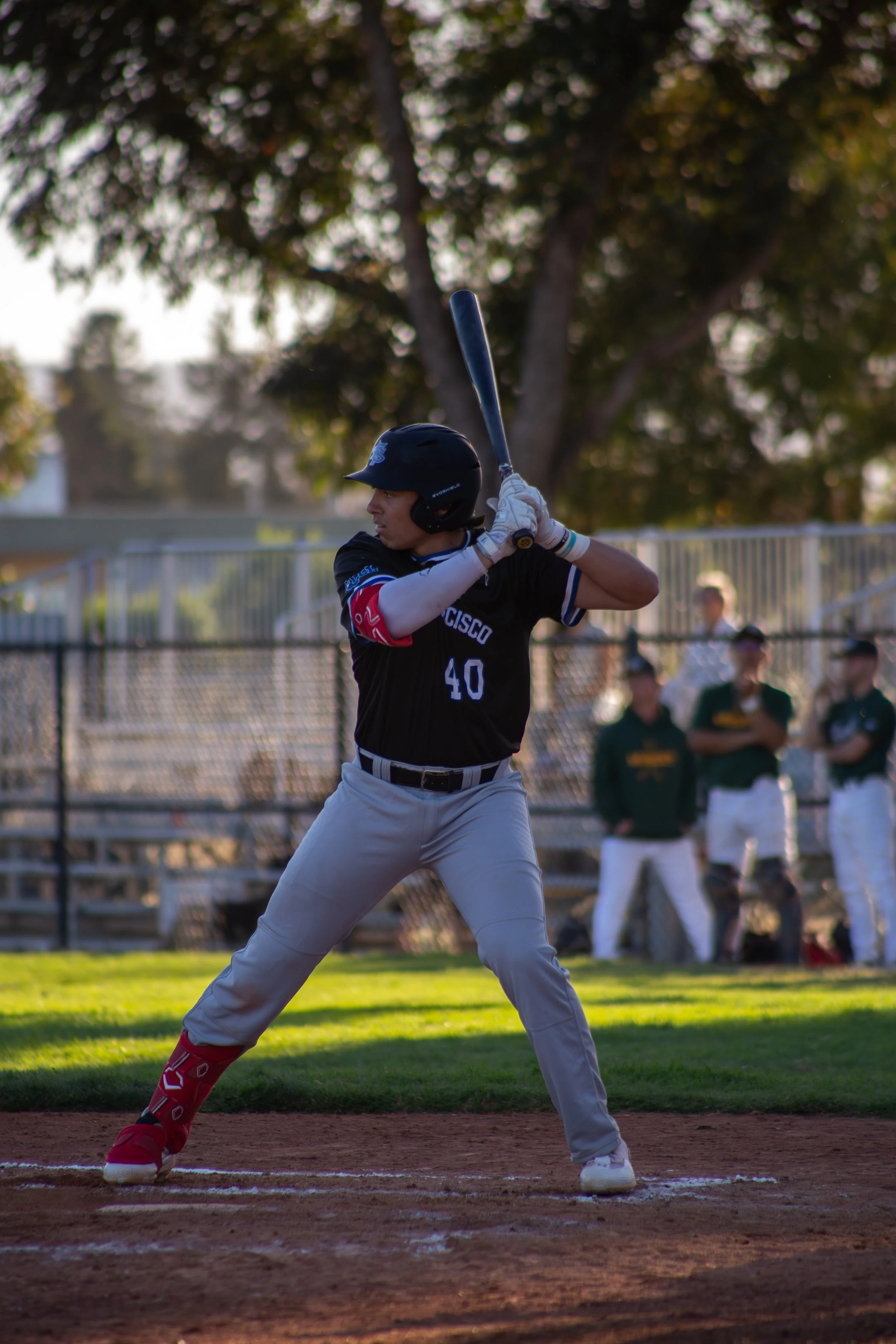 A baseball player at bat during a game, wearing a black helmet and uniform, with onlookers in the background and trees behind the field.