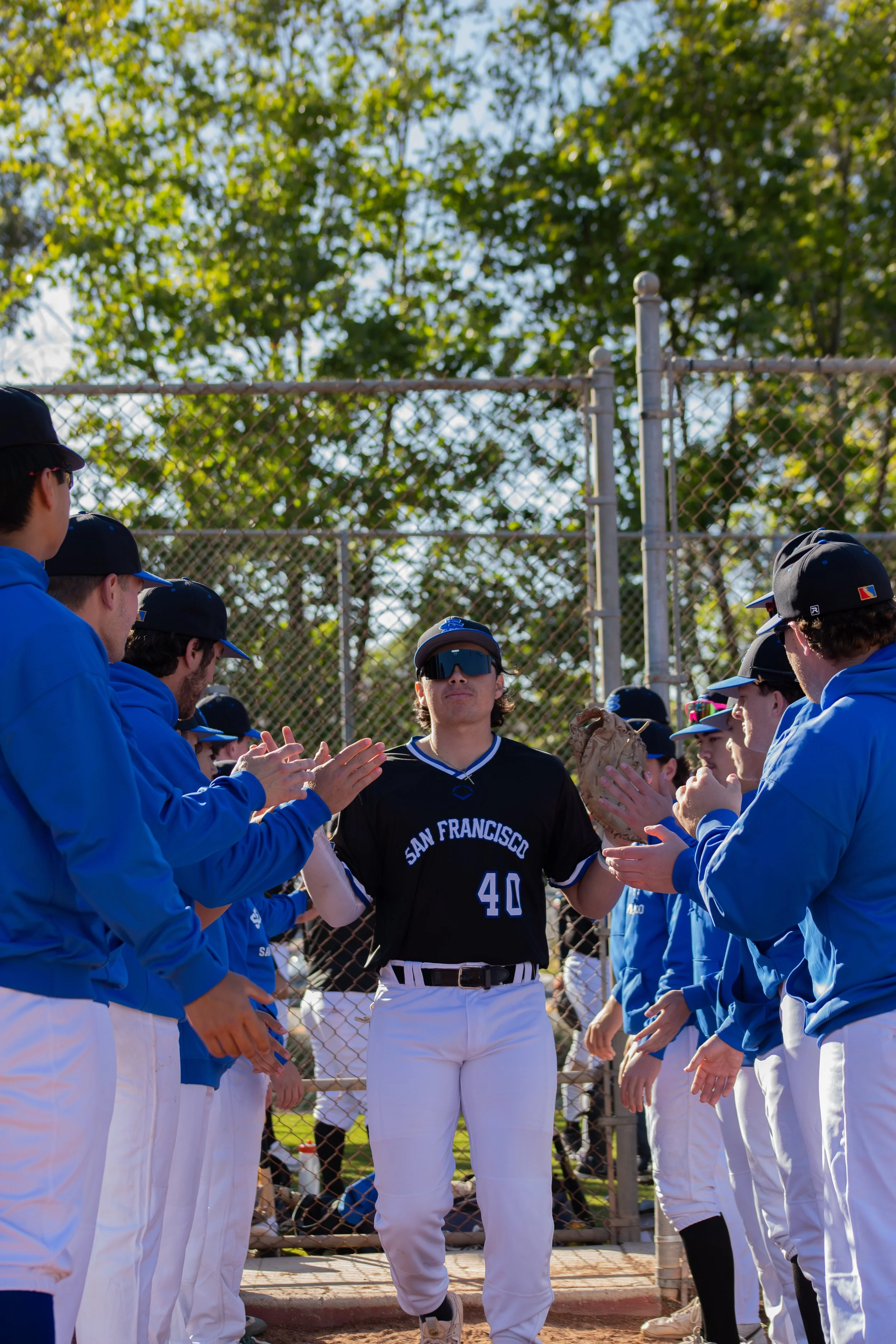 A baseball player in a black uniform with the number 40 and 'San Francisco' written on it stands in the center of a group of players. They are wearing blue jackets, white pants, and black caps, gathered in a line near a chain-link fence. The scene is