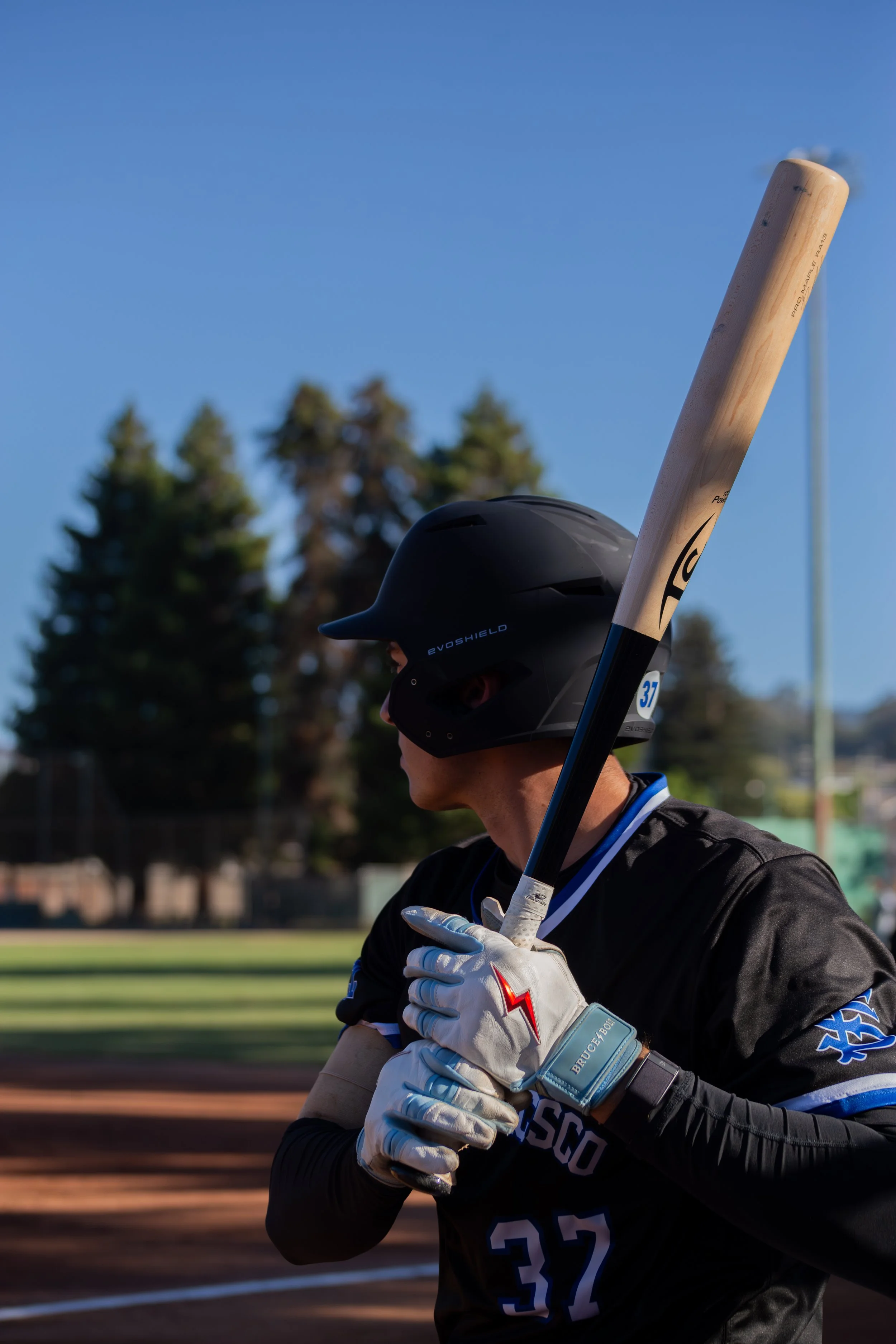 A baseball player wearing a black uniform, a black helmet, and white gloves is holding a bat and walking on a baseball field. There are trees and a chain-link fence in the background.