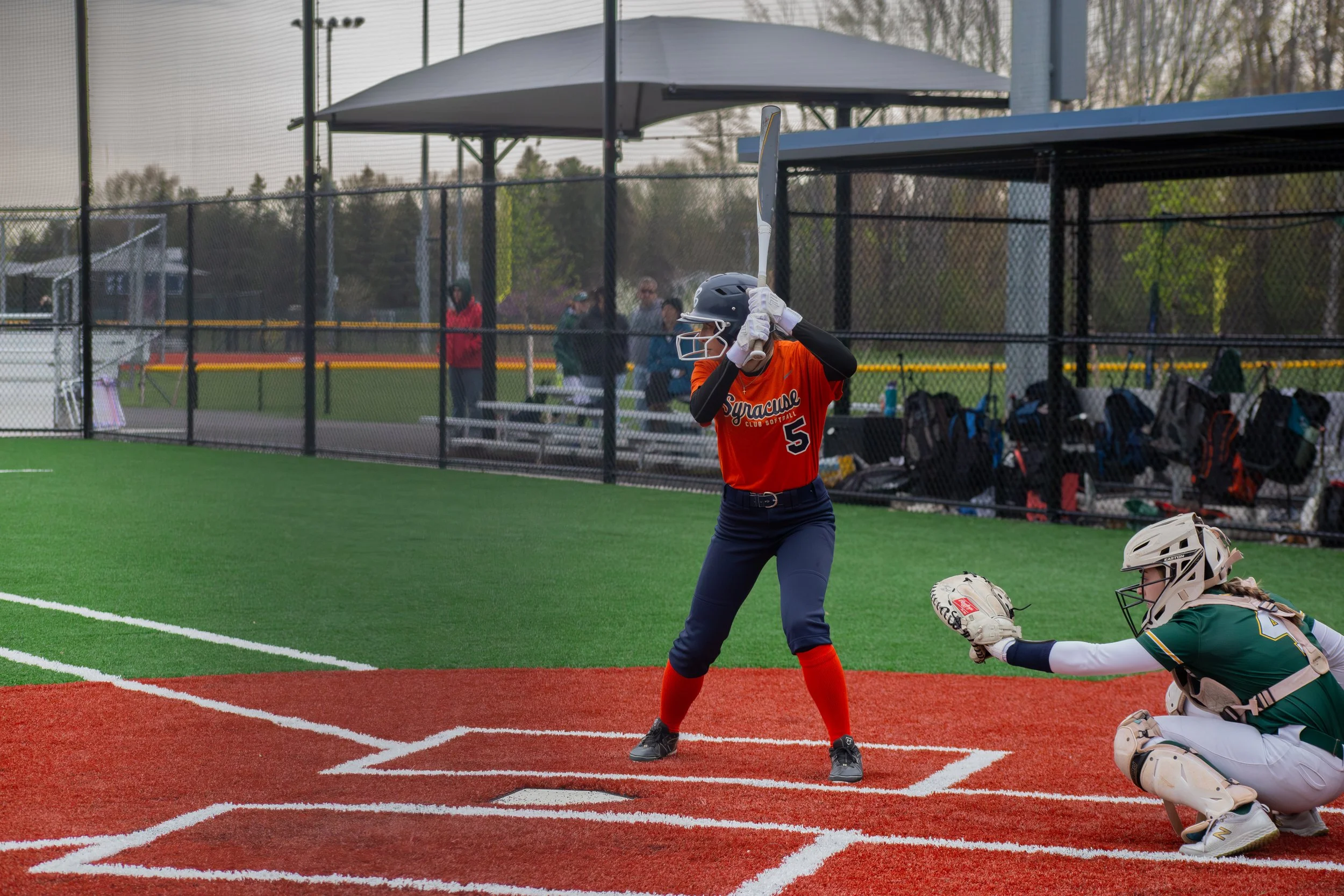 A female softball player stands at the plate with a bat, ready to swing during a game on an outdoor field. She is wearing an orange jersey with the number 5, navy pants, and a black helmet. A female catcher in a green jersey and white pants is crouch