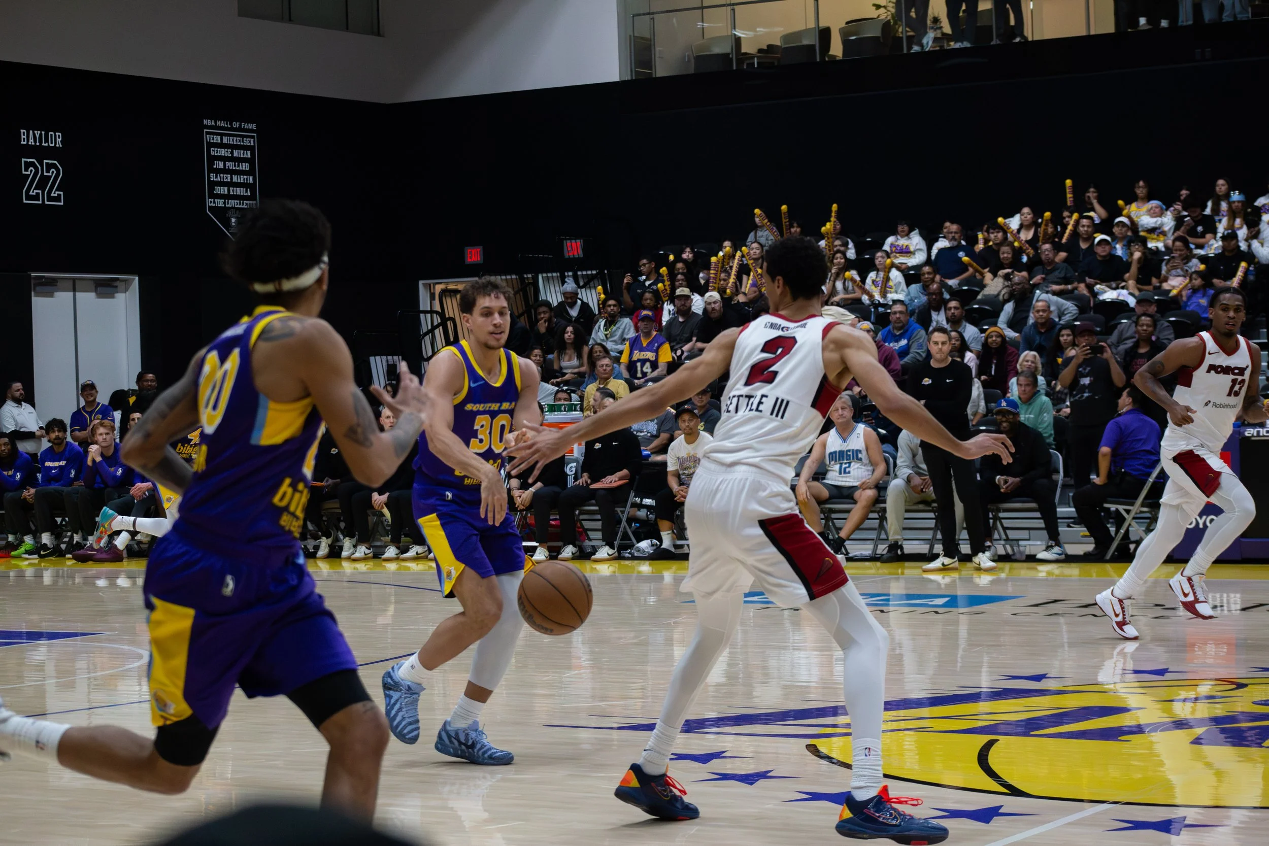A basketball game with players from different teams on an indoor court. Fans sit in the background, some holding foam fingers, and the court features a colorful logo.